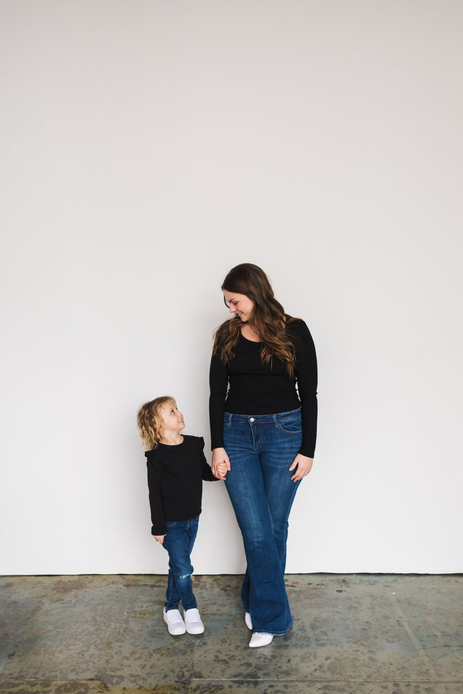 Parent and child stand together against white wall wearing matching black tops and denim.