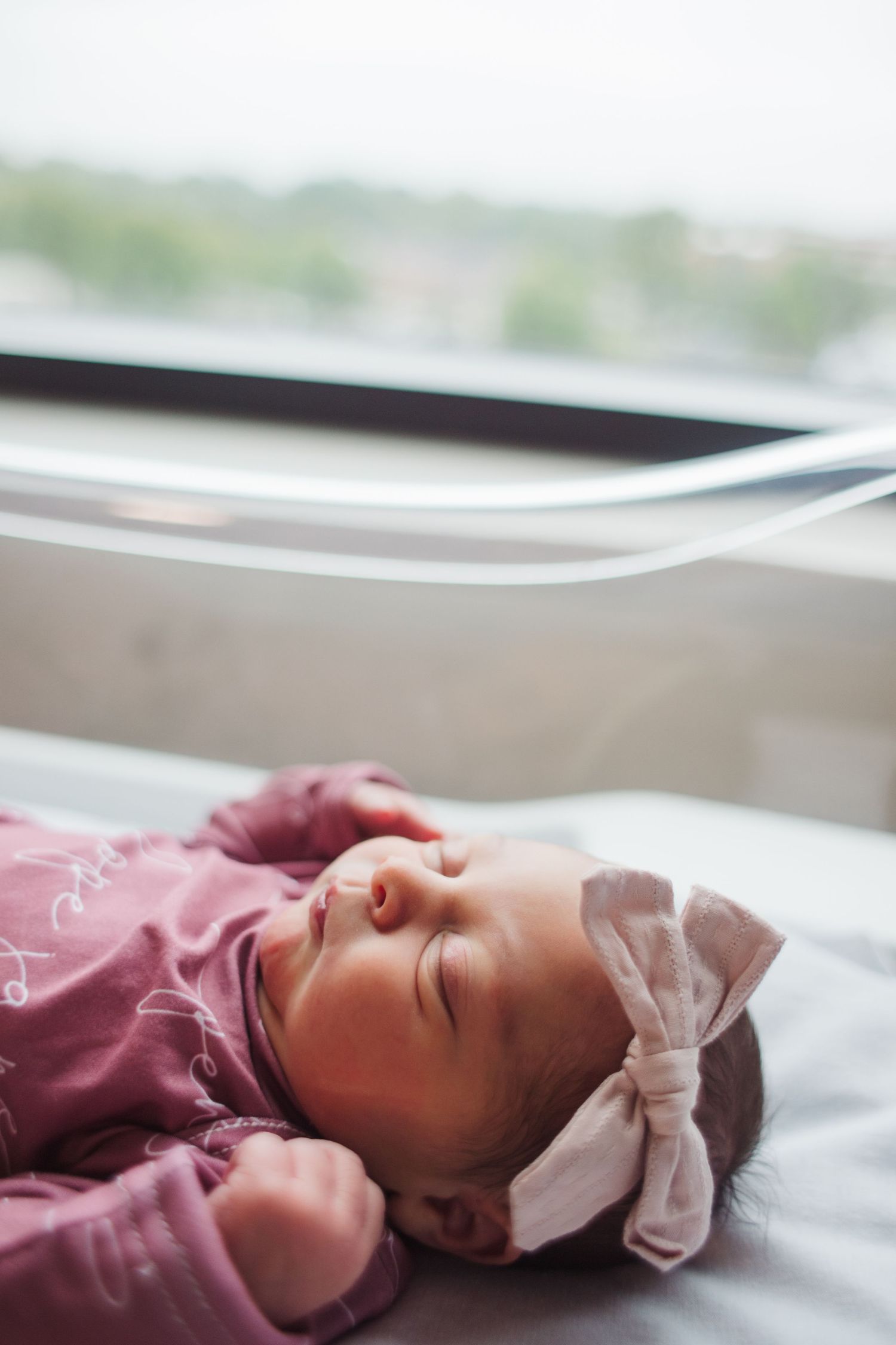 Newborn baby wearing a burgundy outfit with white bow headband rests peacefully on white bedding near window.