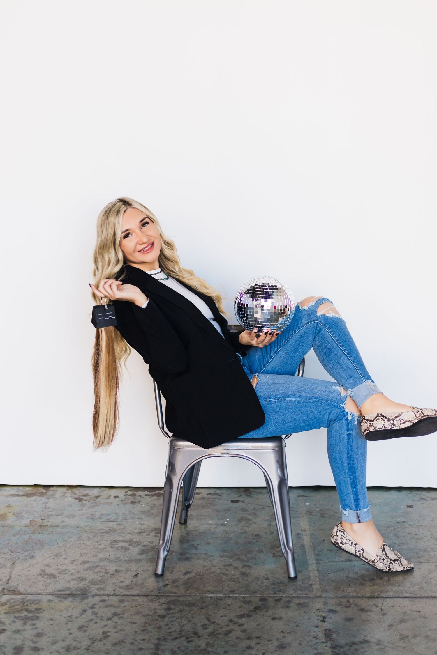 Relaxed pose with disco ball wearing black blazer and ripped jeans against white background.