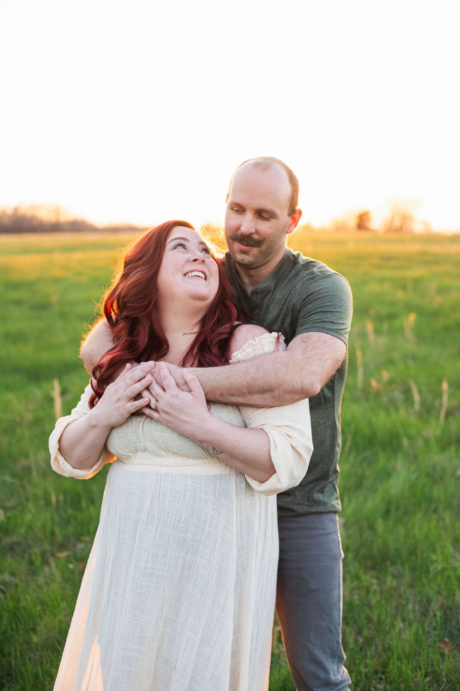 Playful couple embracing in sunlit field during maternity photoshoot.