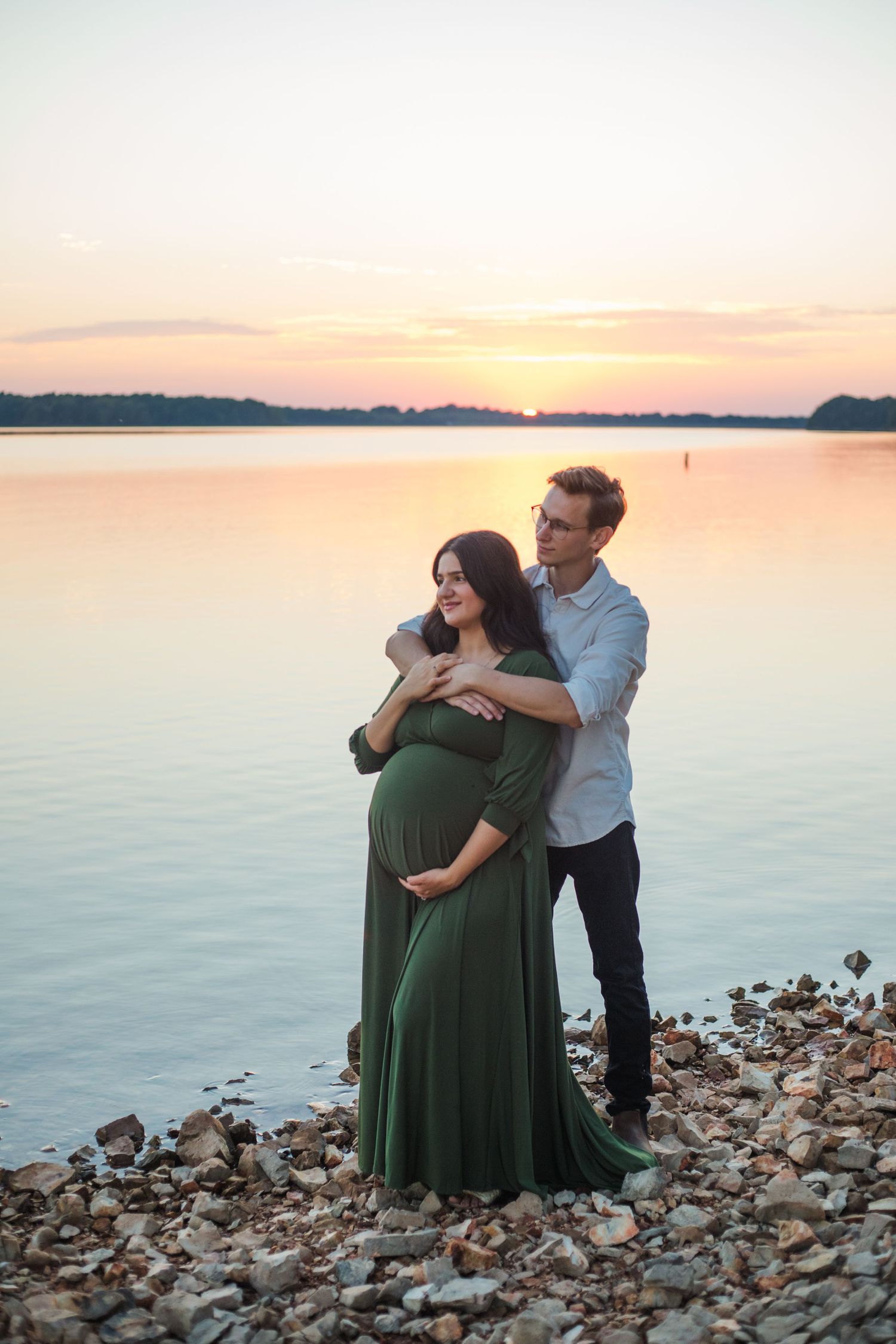A couple embraces on a rocky shoreline at sunset as the golden light reflects off the calm water.