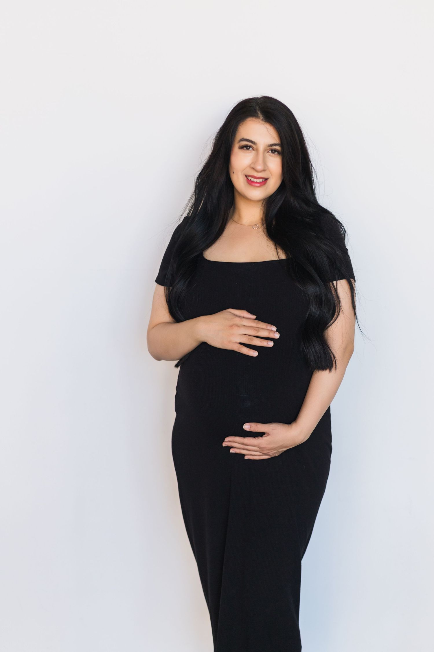 A woman in a black dress cradles her pregnant belly against a light background.