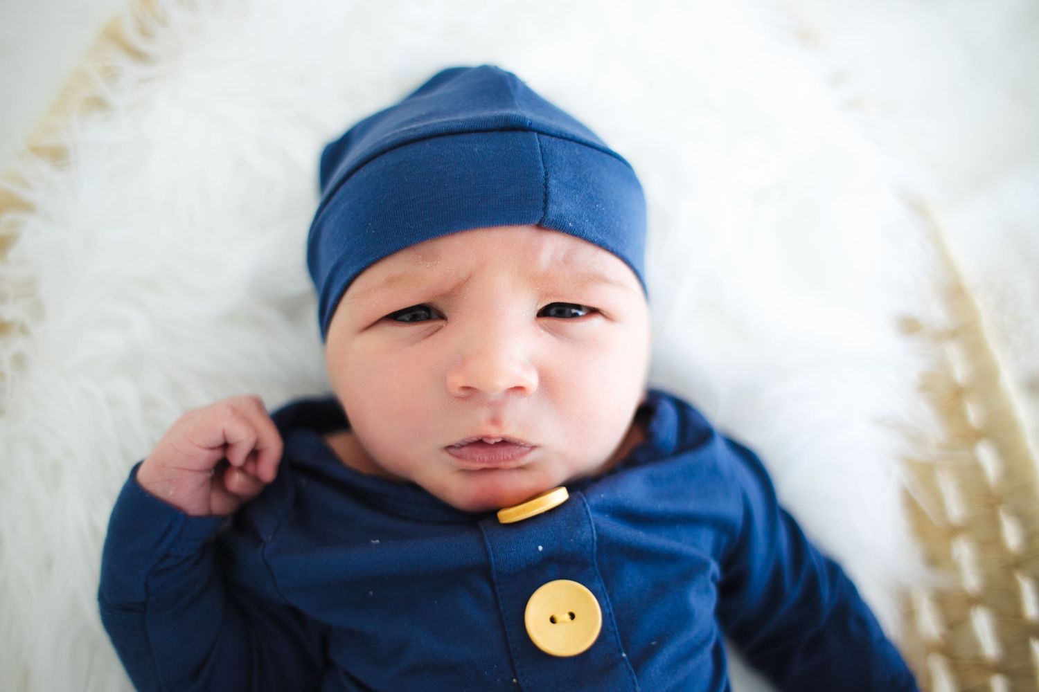 Baby wearing navy blue outfit with matching hat lies on white blanket with decorative wheat stalks in background.