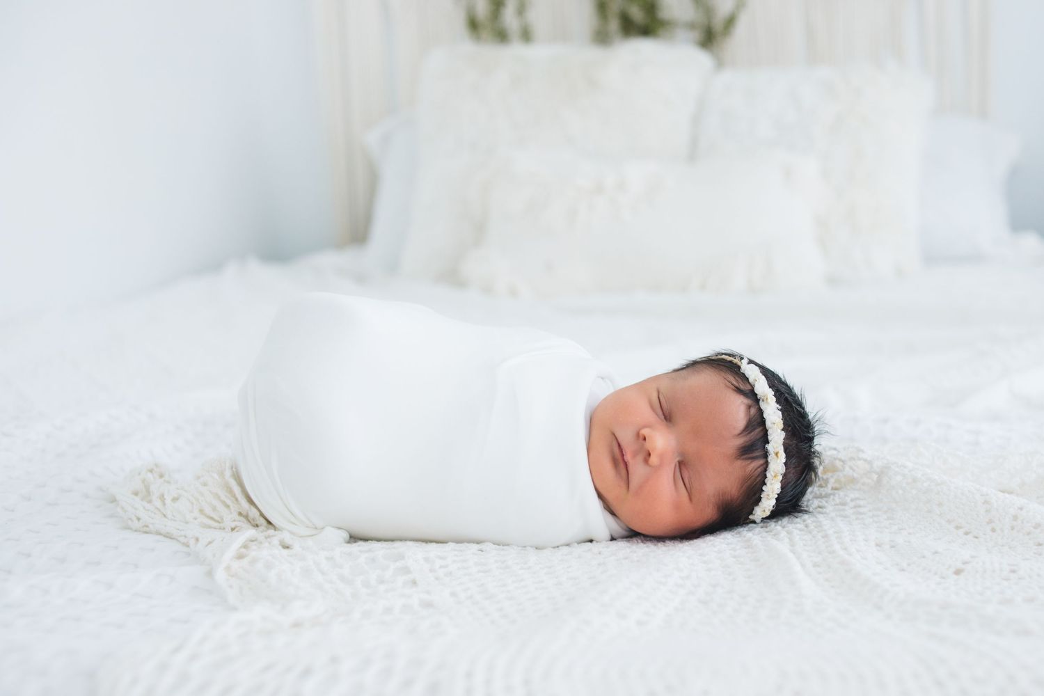 Newborn baby wrapped in white swaddle wearing dark headband sleeps peacefully on white bedding.