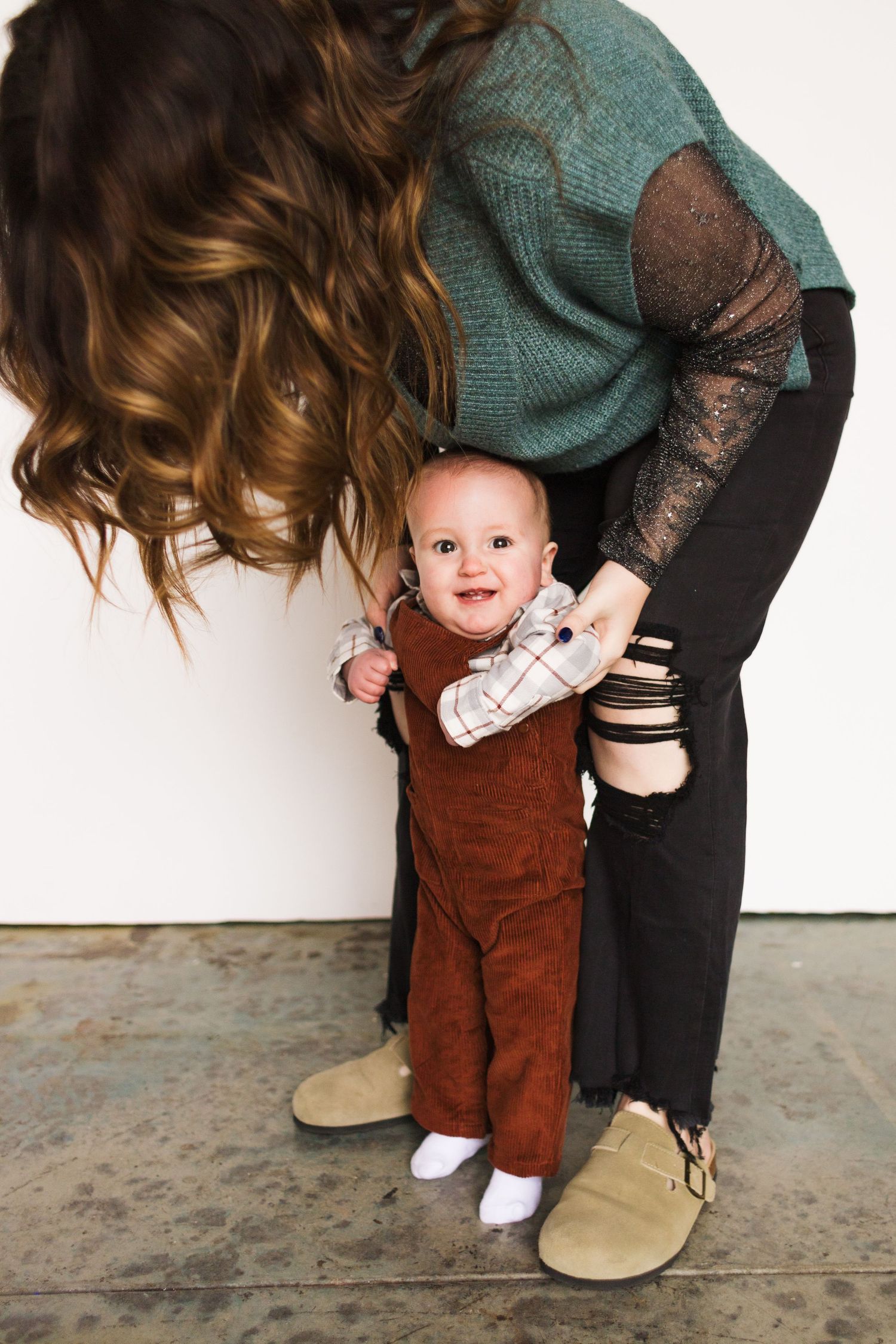 Series showing parent bending down to help baby in brown overalls learn to stand.