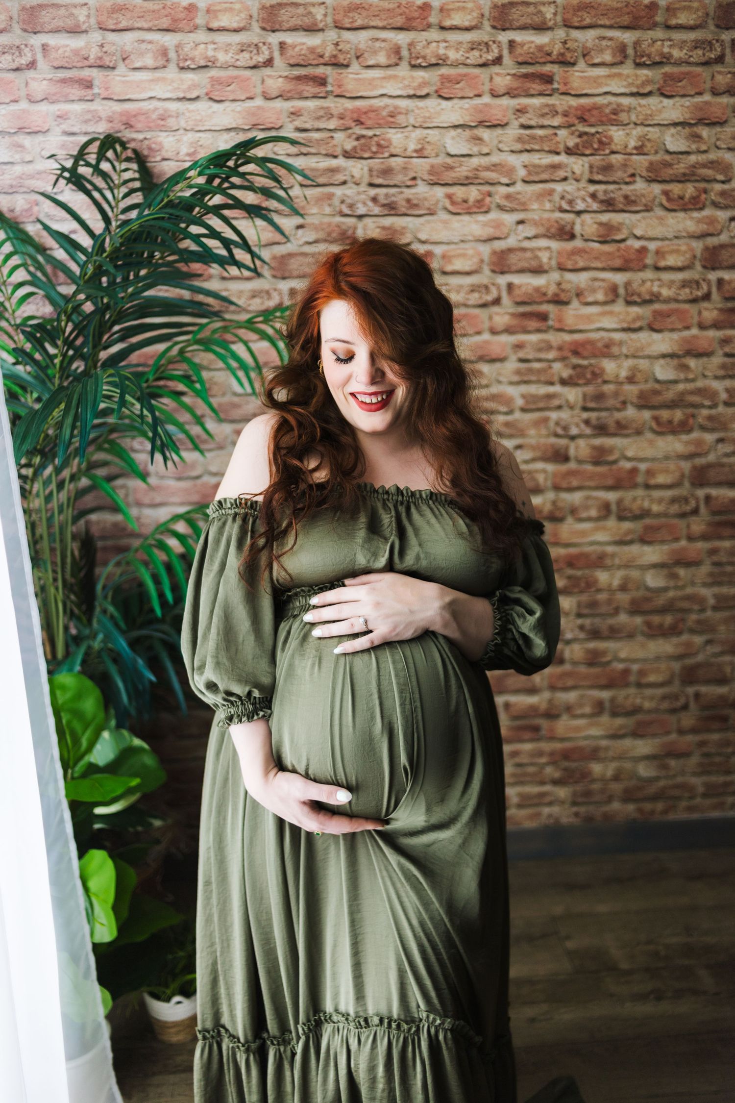 Maternity portrait in olive green dress against brick wall with tropical plants.