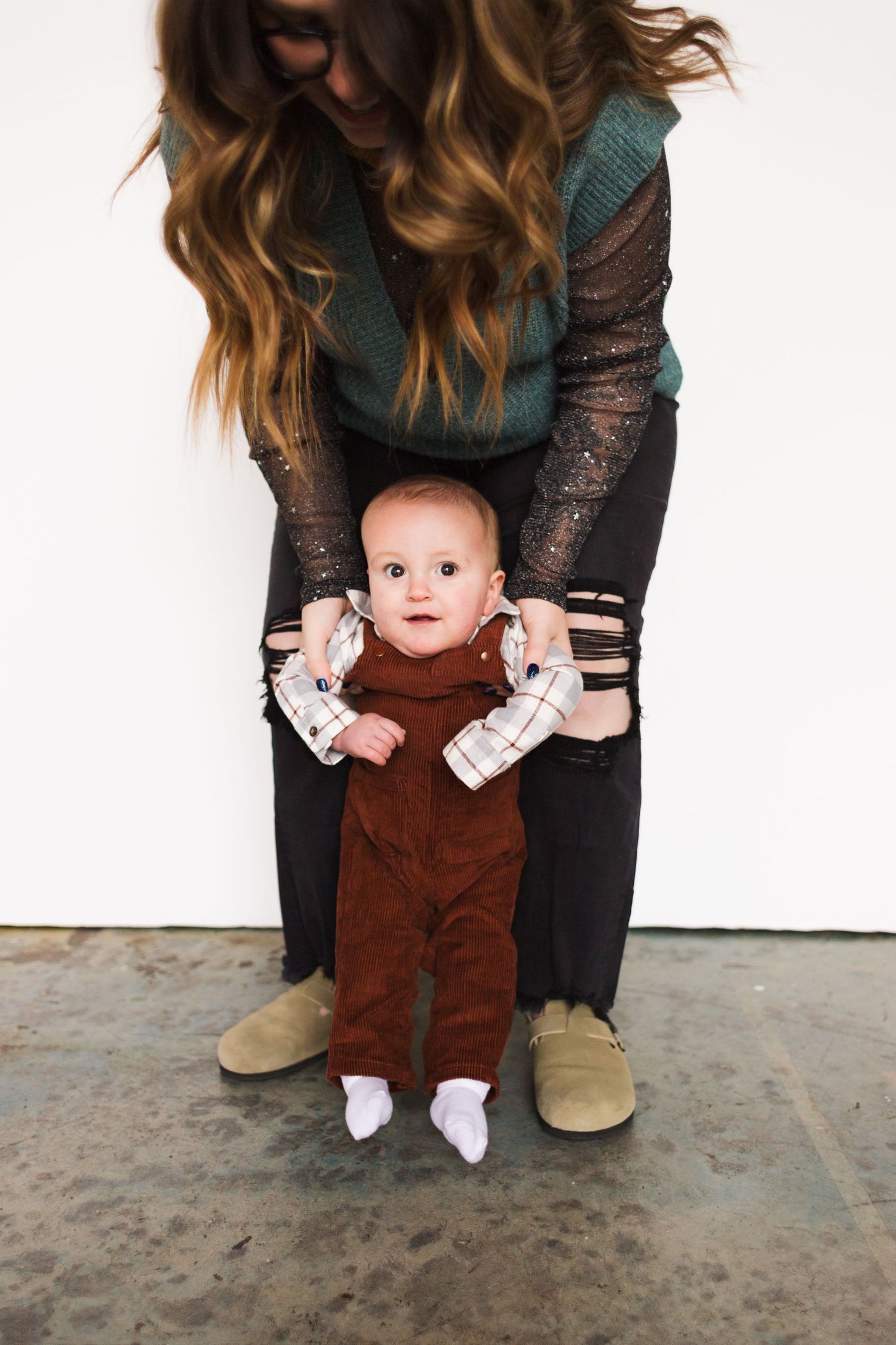 Parent helps baby in brown overalls practice standing on concrete floor.