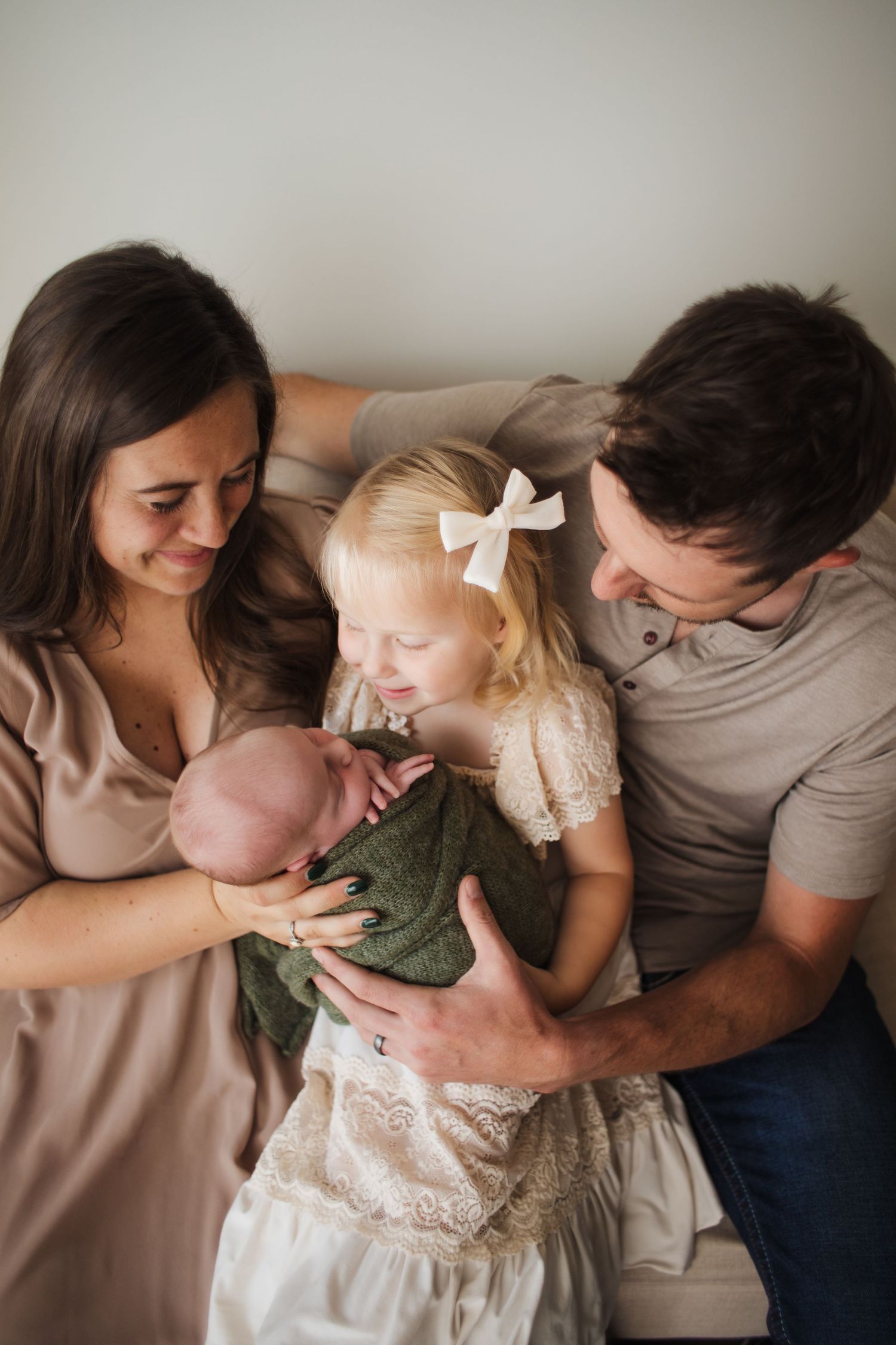 Family of four with parents and toddler girl admiring their newborn baby wrapped in green blanket.