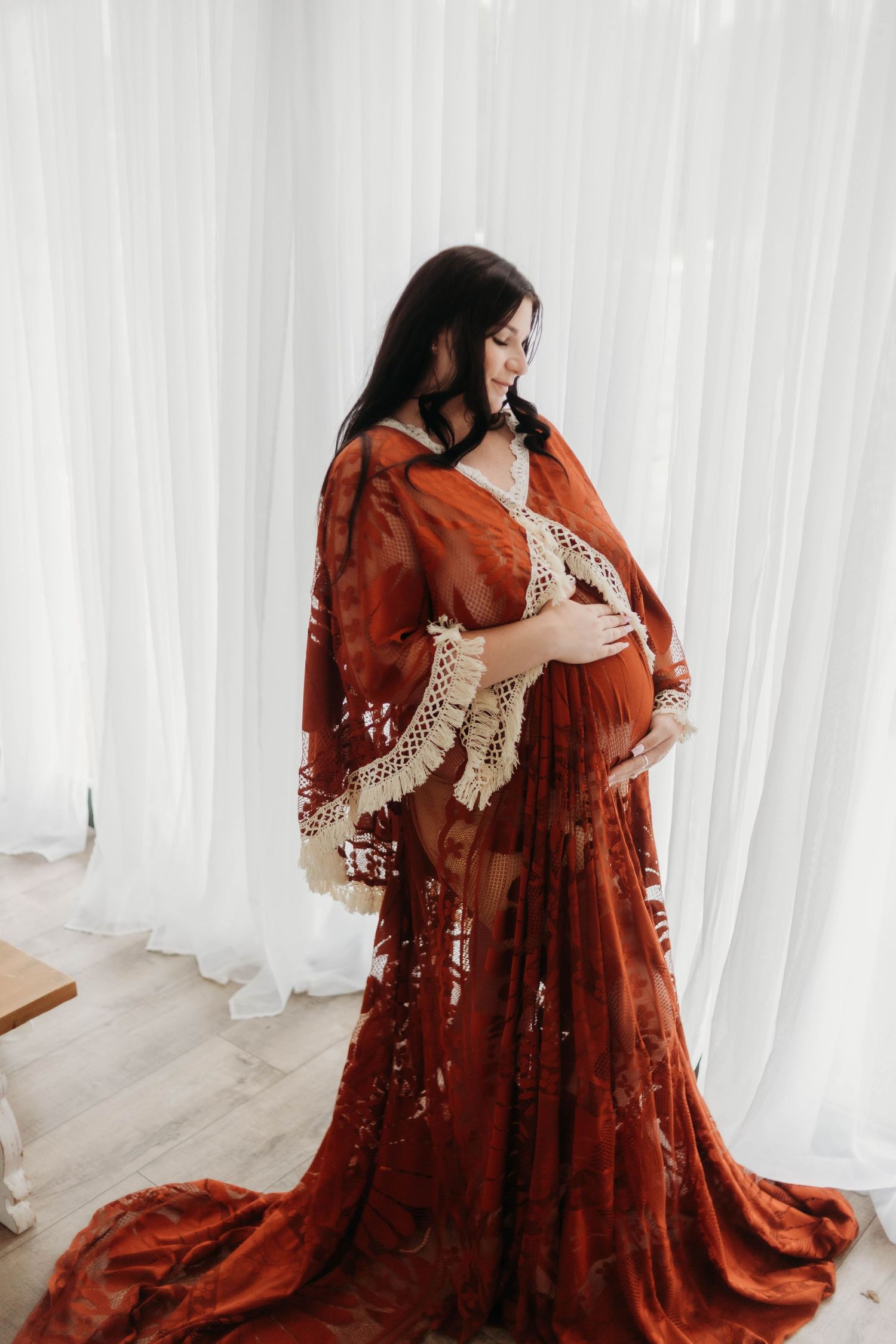 Dramatic maternity portrait in flowing rust-colored lace gown against white backdrop.