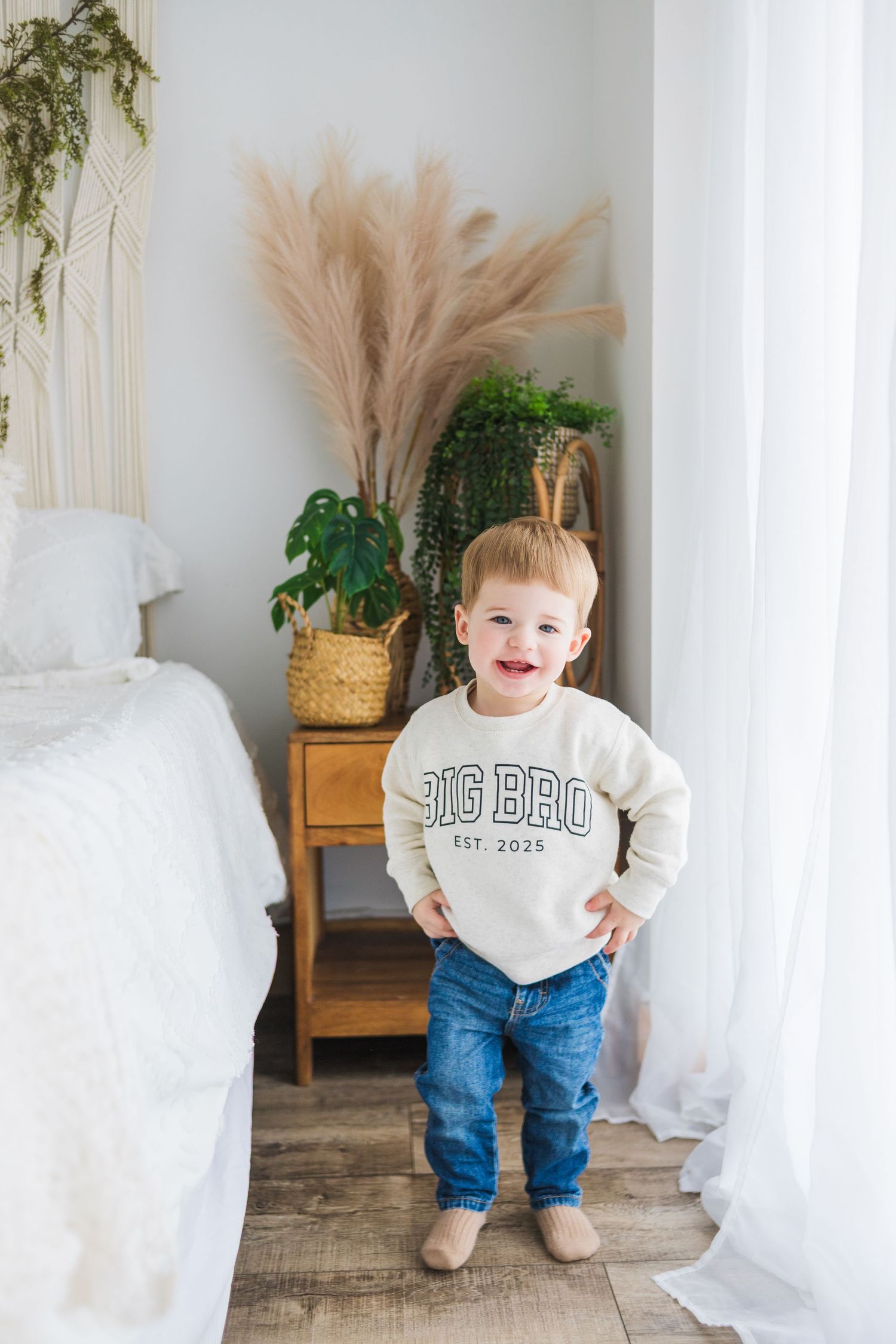 Toddler in Big Bro sweatshirt and jeans stands between white curtains near decorative plants.