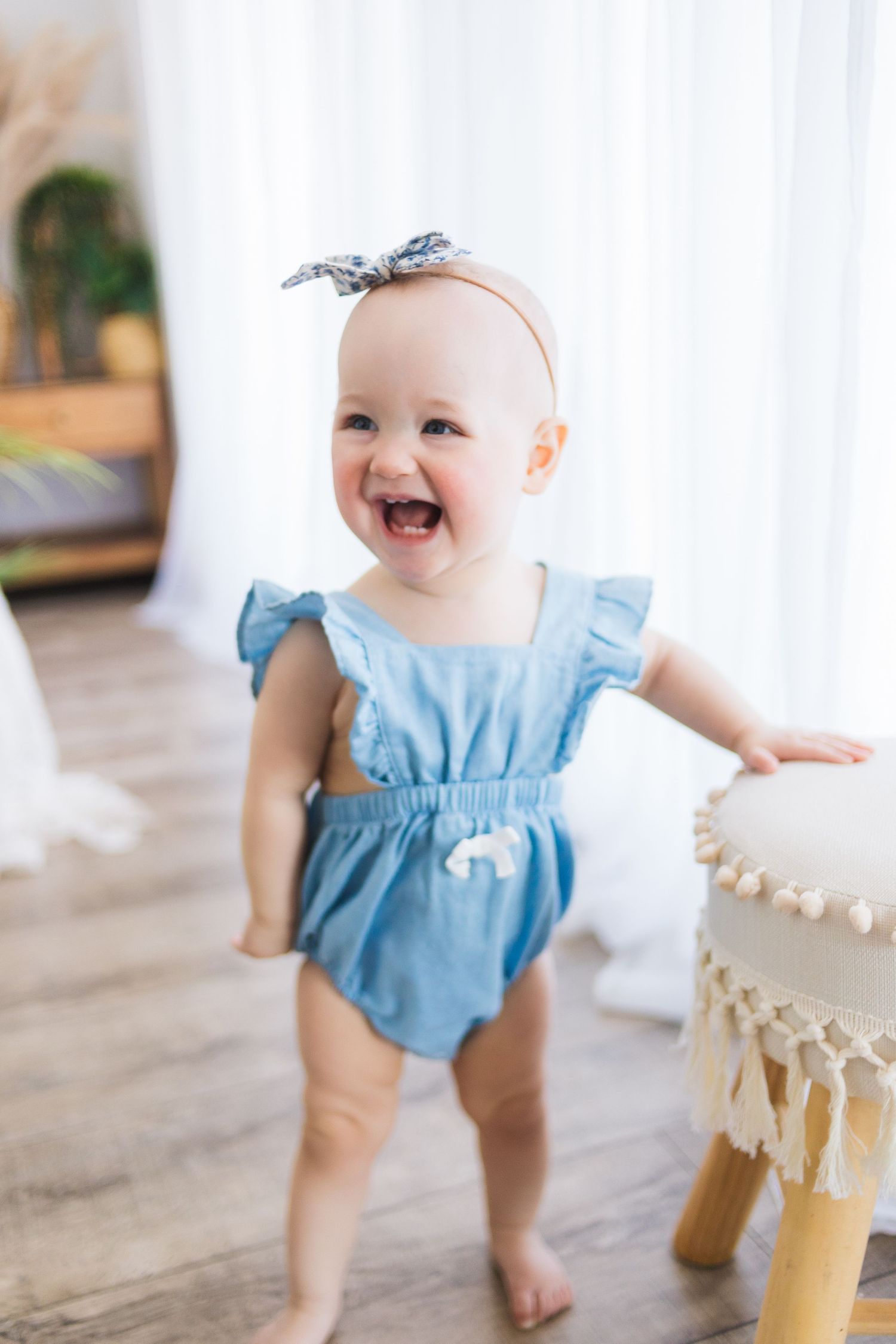 Baby wearing light blue romper takes first steps while holding onto macrame chair in bright sunlit room.