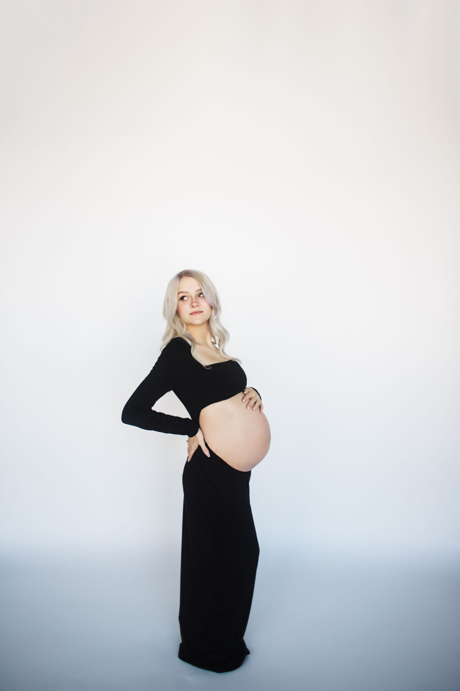 Expectant mother in long black dress poses against white background showing pregnant belly in elegant maternity photo.