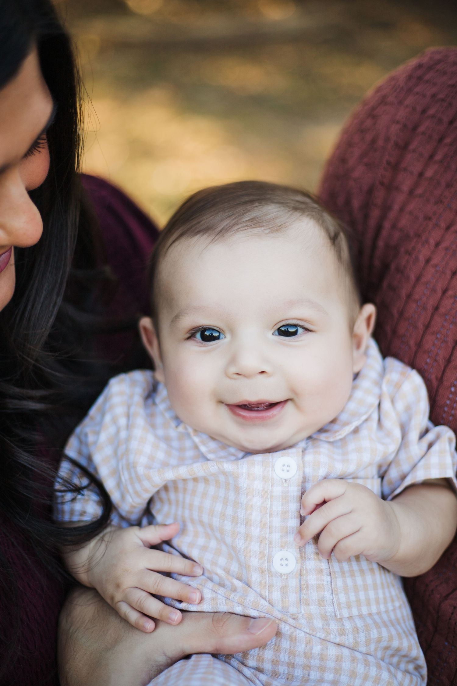 A baby in a light checkered outfit smiles while being held, with bright blue eyes and a joyful expression.