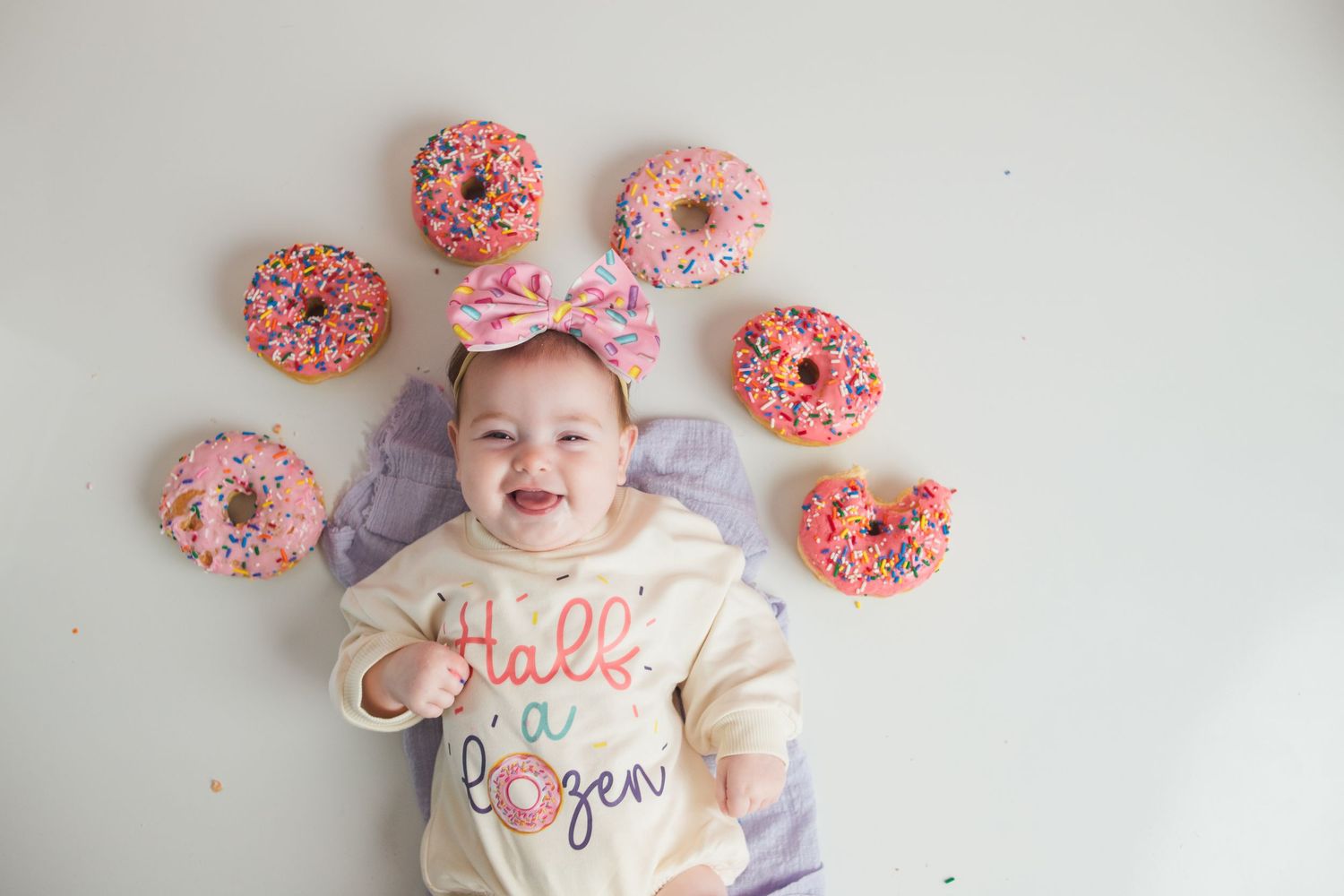 Baby lying on white blanket surrounded by pink frosted donuts in a styled photography setup.