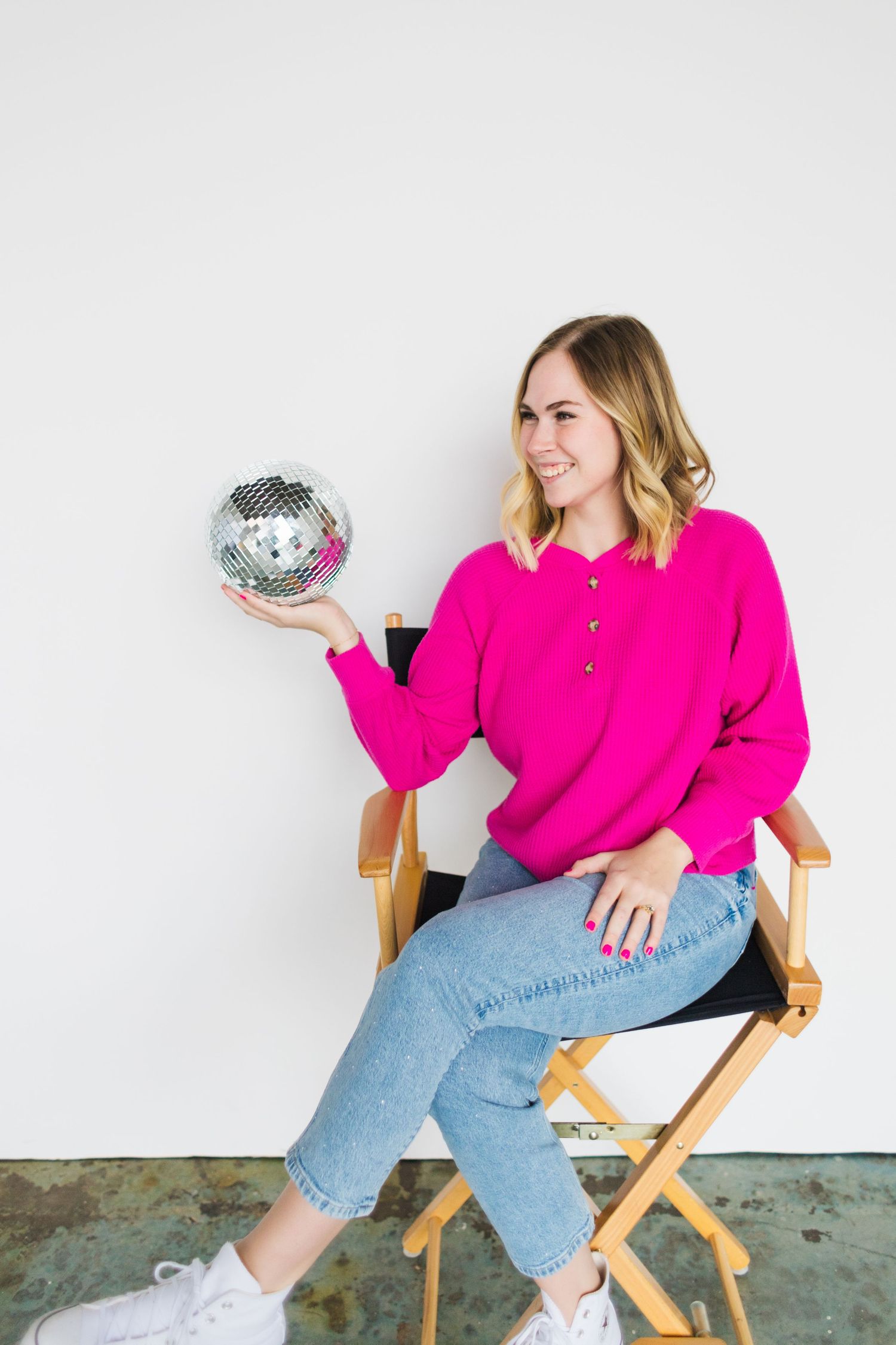 Person in hot pink sweater holds disco ball while sitting in director's chair against white backdrop.