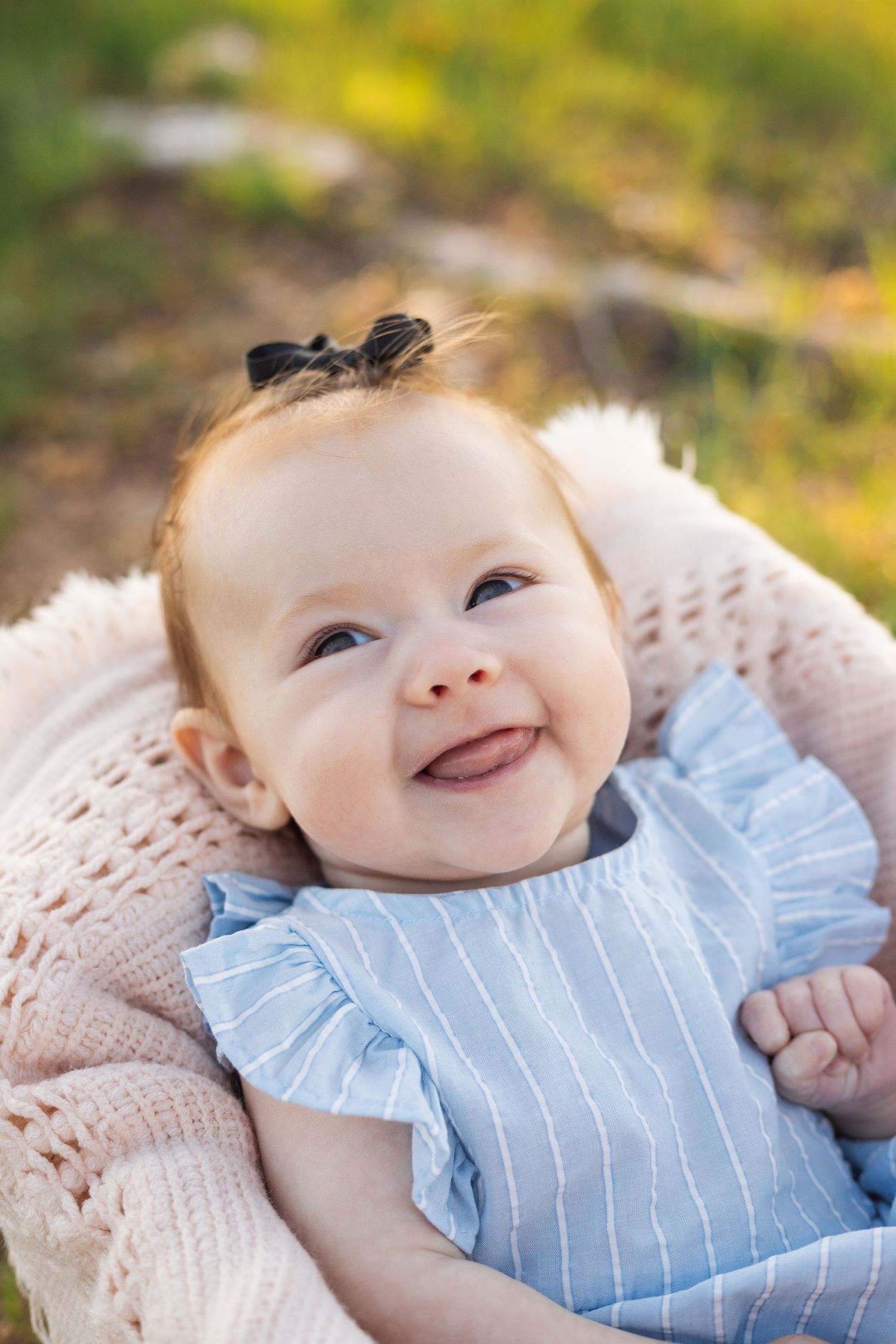 A baby wearing a light blue striped outfit smiles while lying on a knitted blanket in outdoor natural lighting.