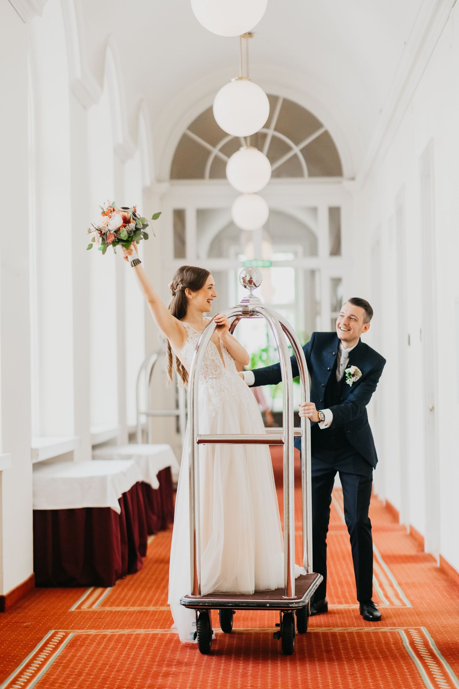 A joyful couple having fun with a luggage cart in an elegant hotel hallway with red carpeting and white walls.