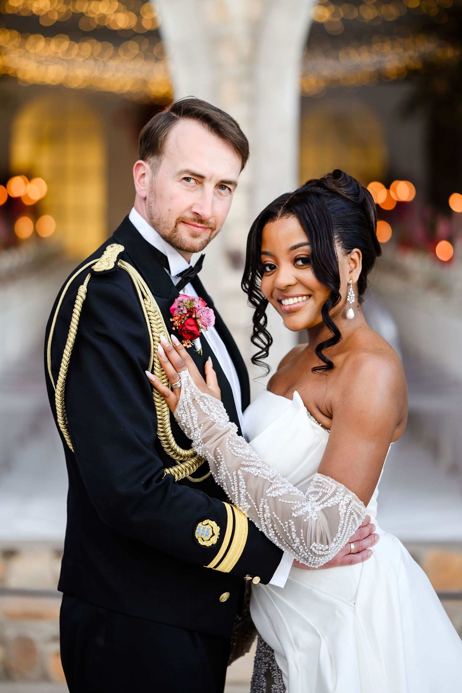A newlywed couple in formal attire embraces during their wedding day under glowing golden lights.