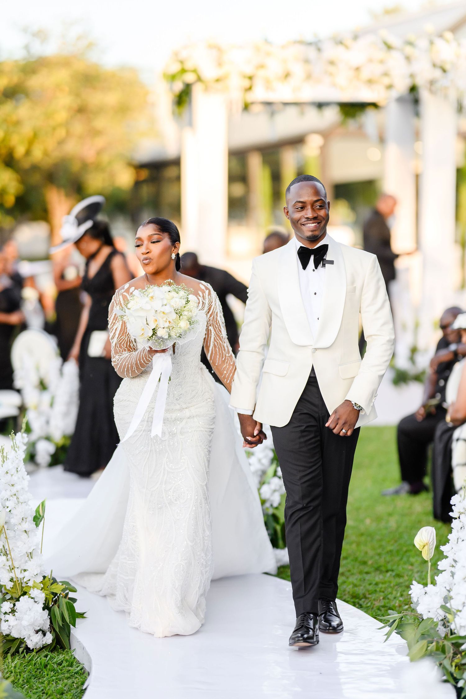 Happy couple walking down white aisle runner decorated with flowers at outdoor wedding ceremony in Lusaka