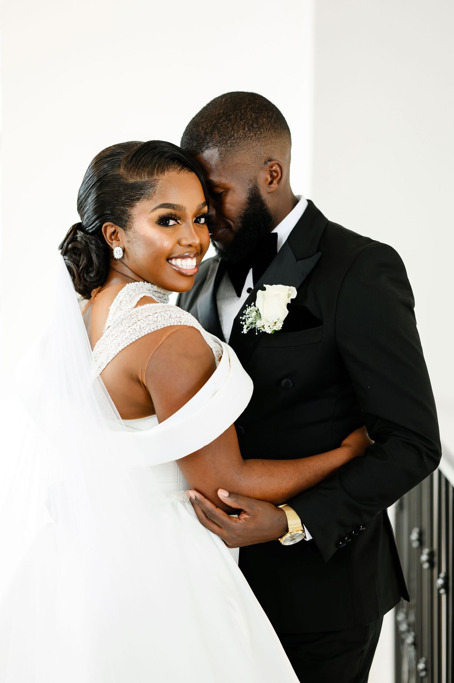 A romantic wedding portrait showing a bride in a white off-shoulder gown and groom in black tuxedo sharing an intimate moment.