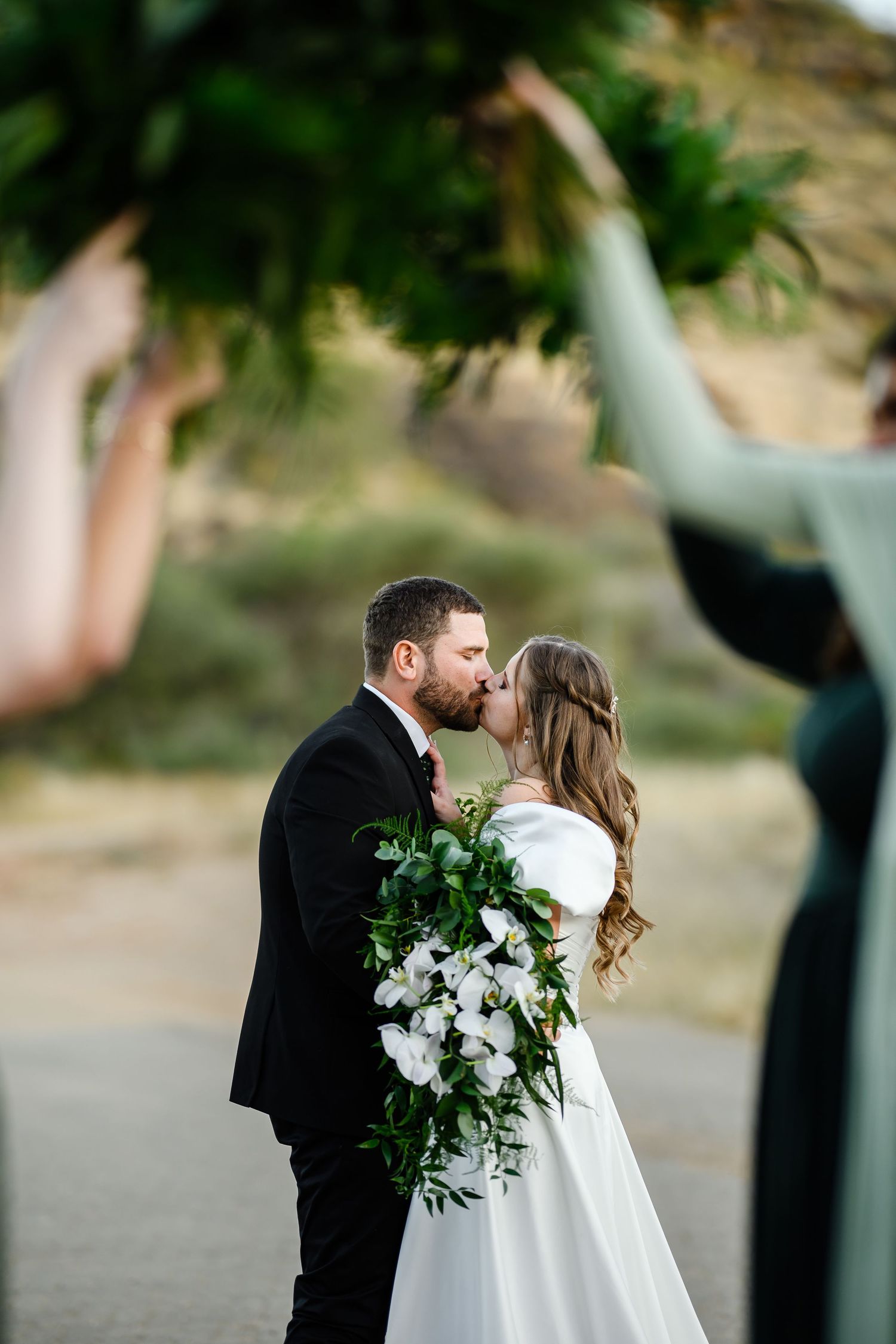 A couple in wedding attire embraces outdoors with the bride holding a large white and green floral bouquet.