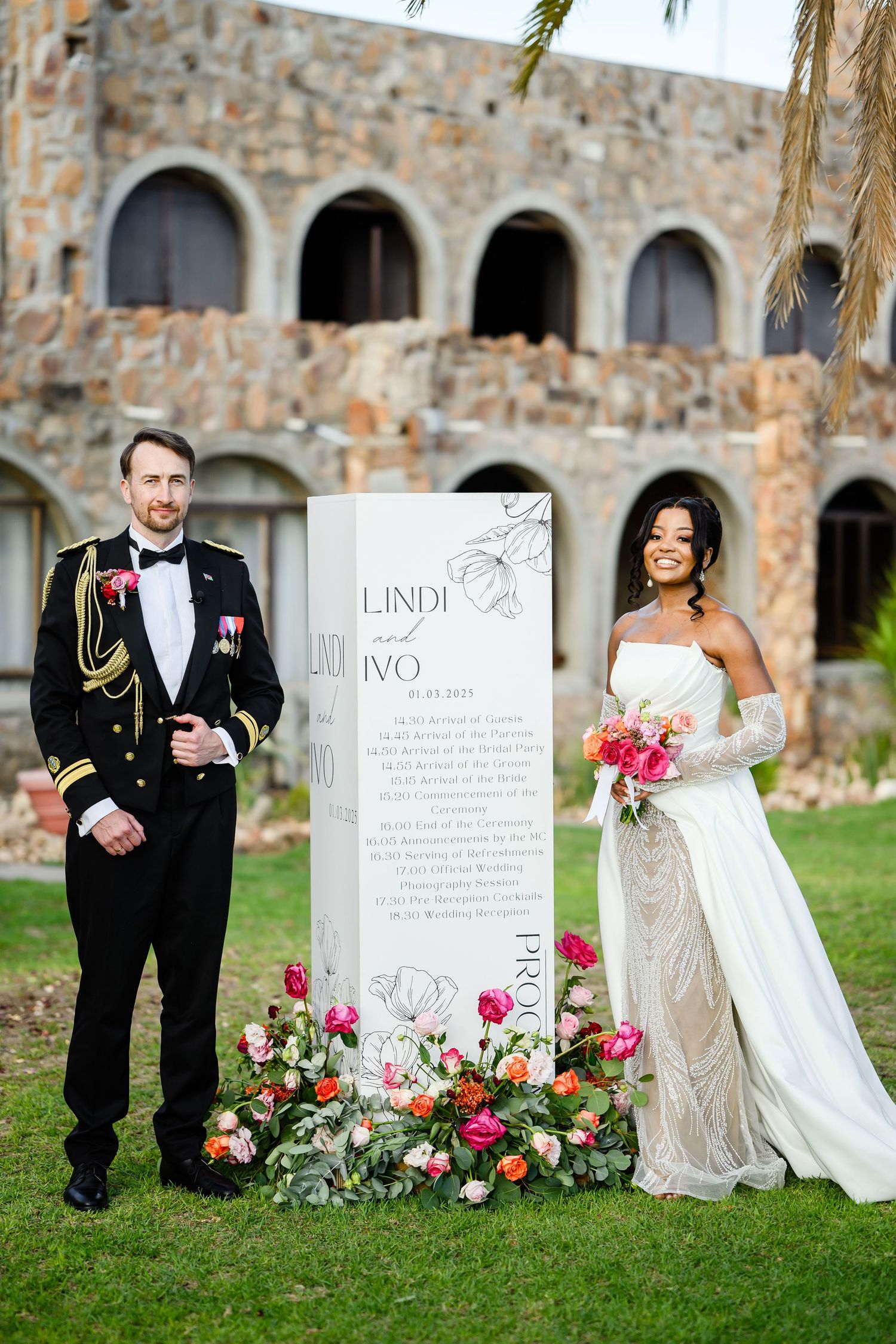 A bride in white and a groom in military dress stand beside an elegant wedding sign with floral arrangements on a lawn.