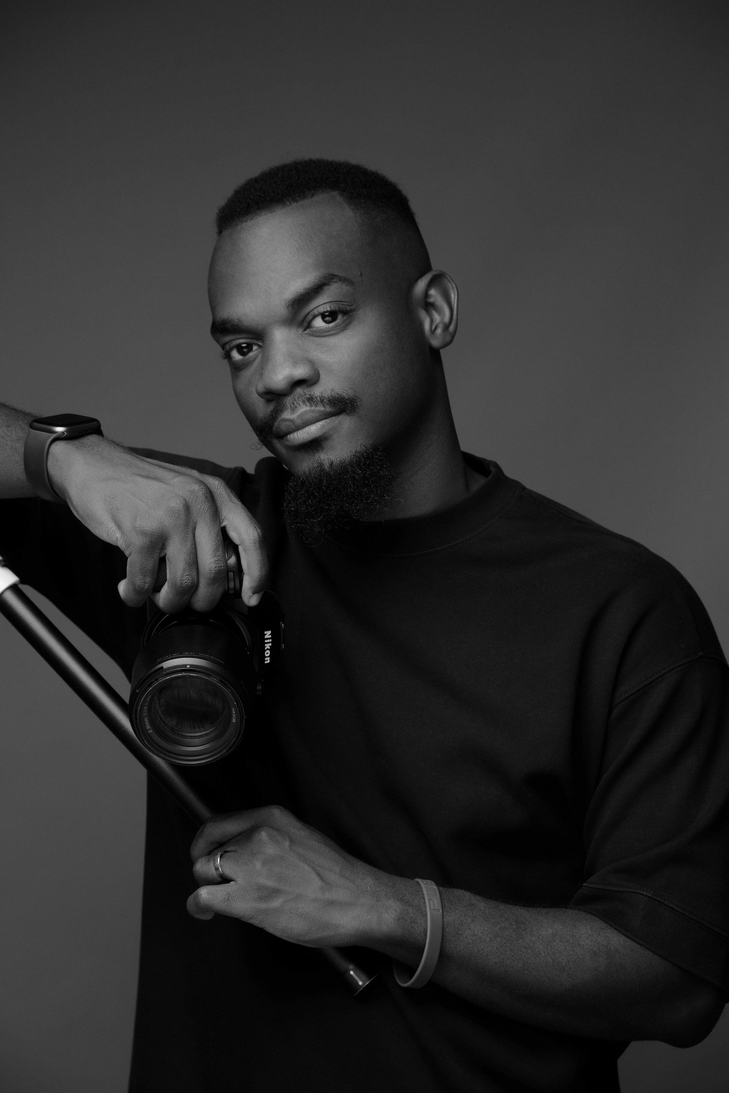 Professional photographer in black sweater posing with camera against gray background.