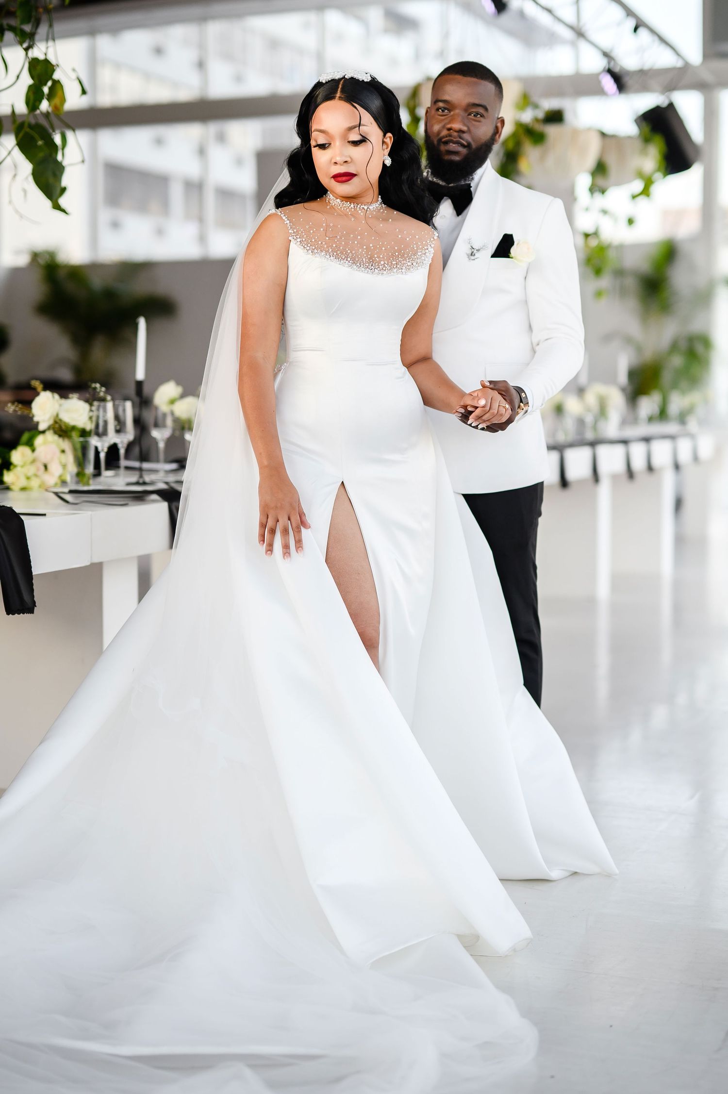 A couple in elegant wedding attire, with a white gown and white tuxedo jacket, pose together in a light-filled venue.
