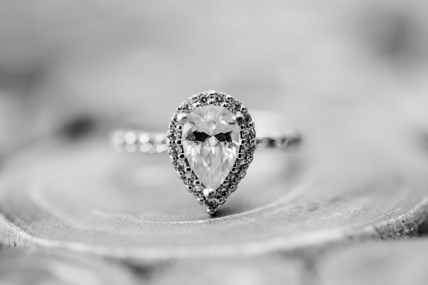 Close-up black and white shot of a pear-shaped diamond engagement ring with halo setting on textured surface.