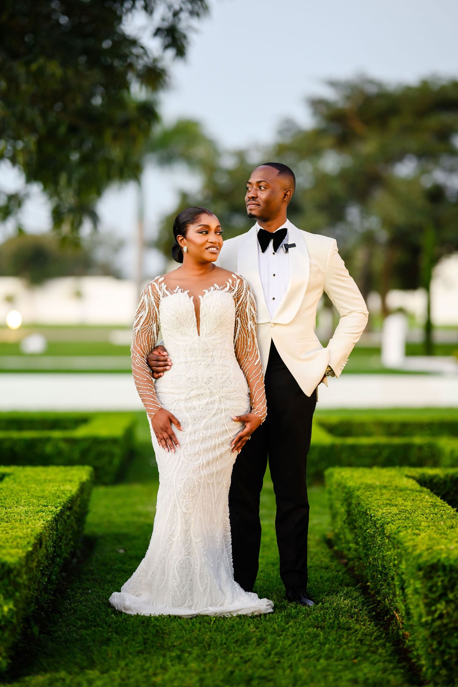 Newlywed couple poses elegantly in a formal garden, with the bride in lace and groom in white dinner jacket.