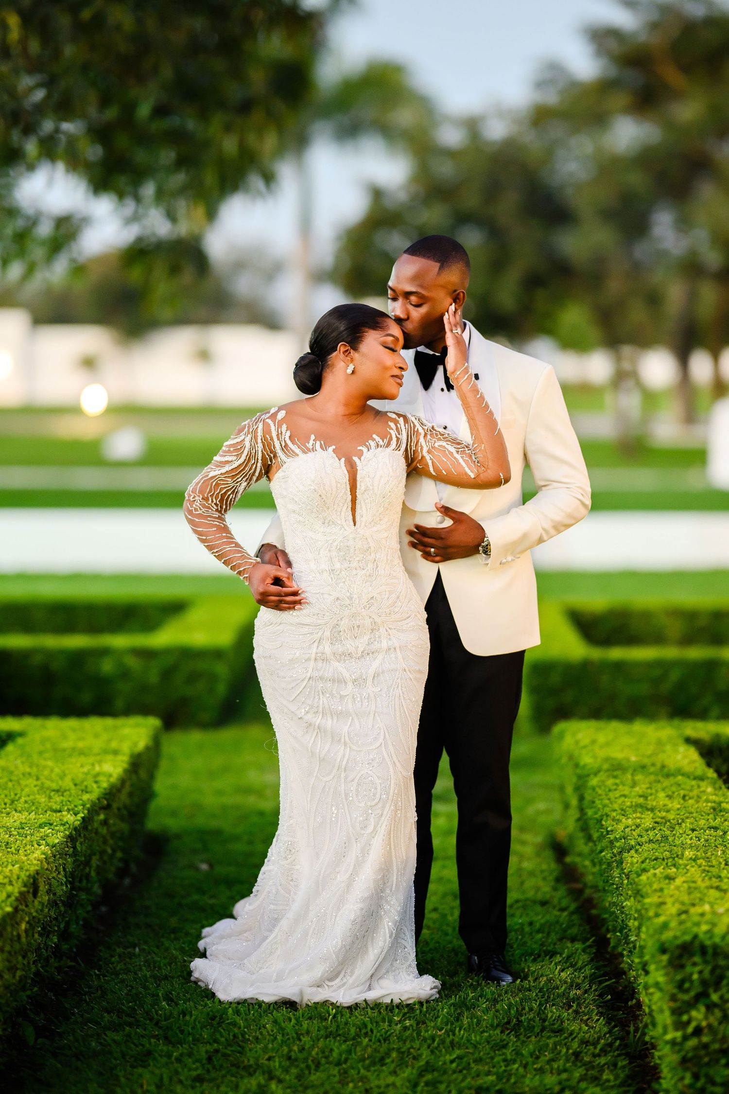 Newlywed couple poses romantically in garden with manicured hedges, bride in lace gown and groom in ivory jacket.