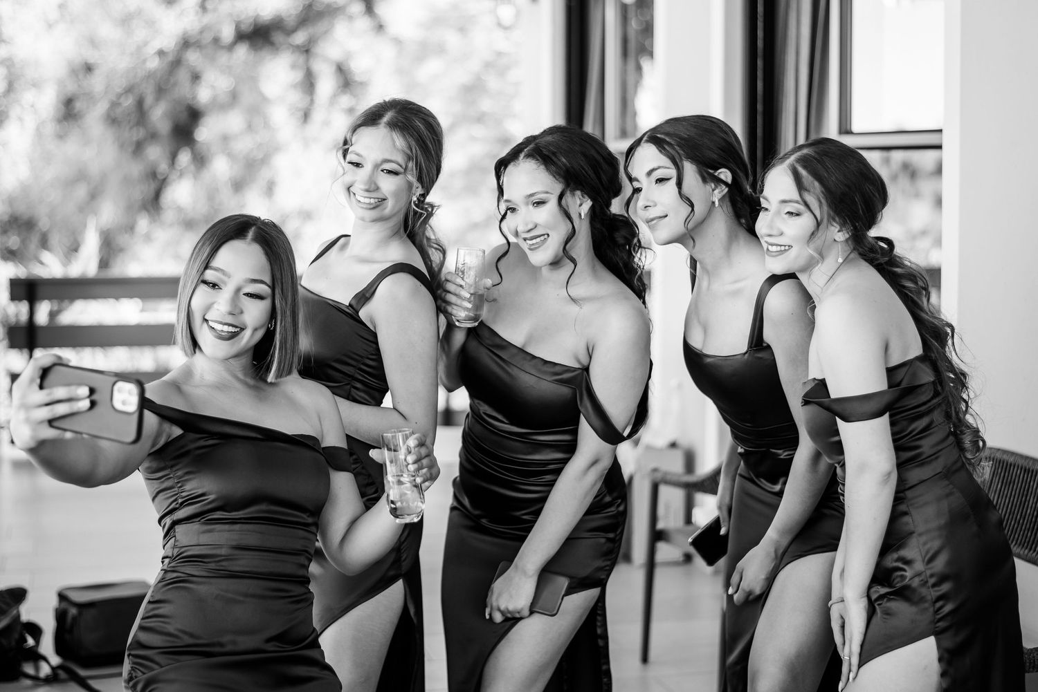 Five bridesmaids in matching black dresses pose together and laugh during a wedding photoshoot.