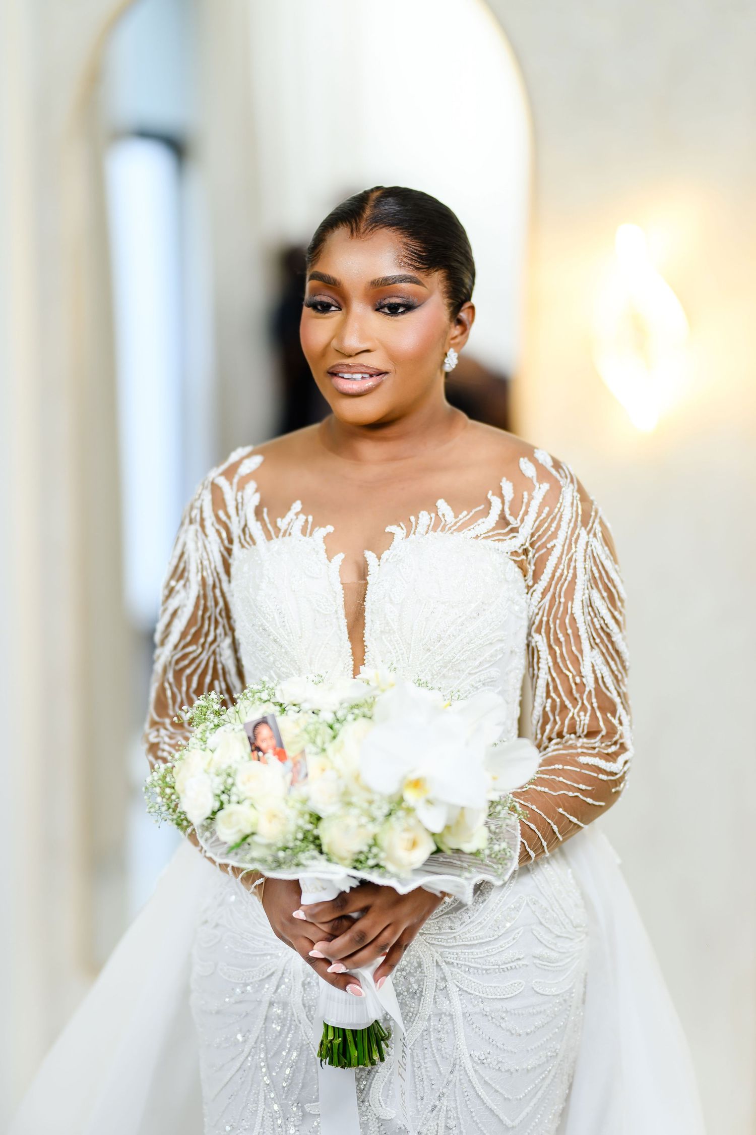 Beautiful bride in long sleeve sheer lace wedding gown holds white and green bouquet against light interior backdrop.