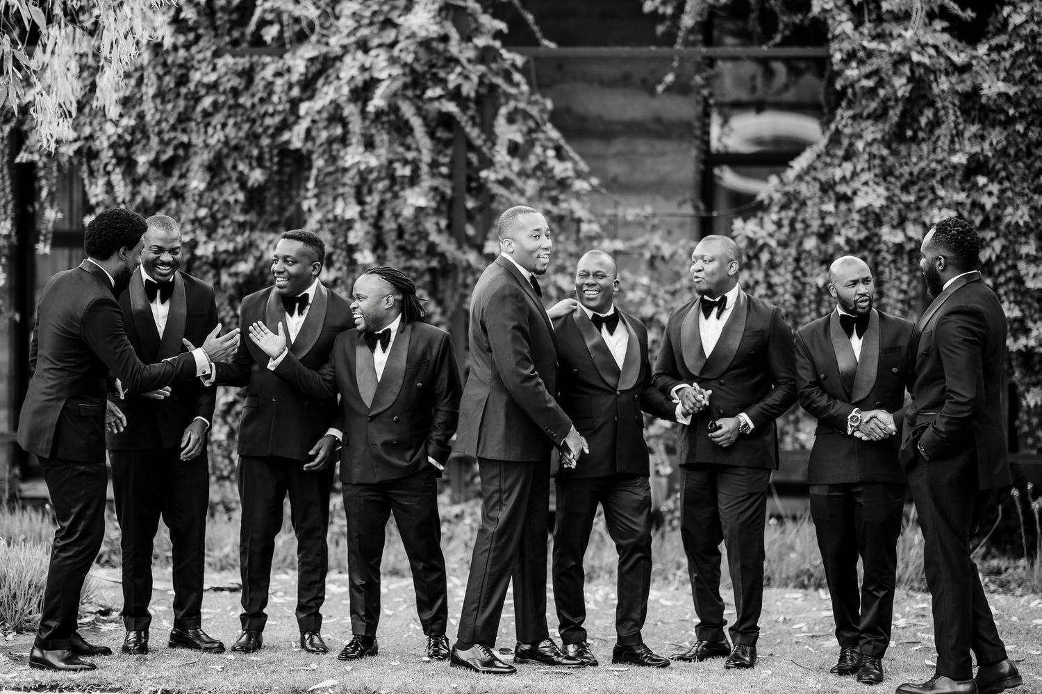 A black and white photo of groomsmen in formal attire standing together outdoors during a wedding celebration.