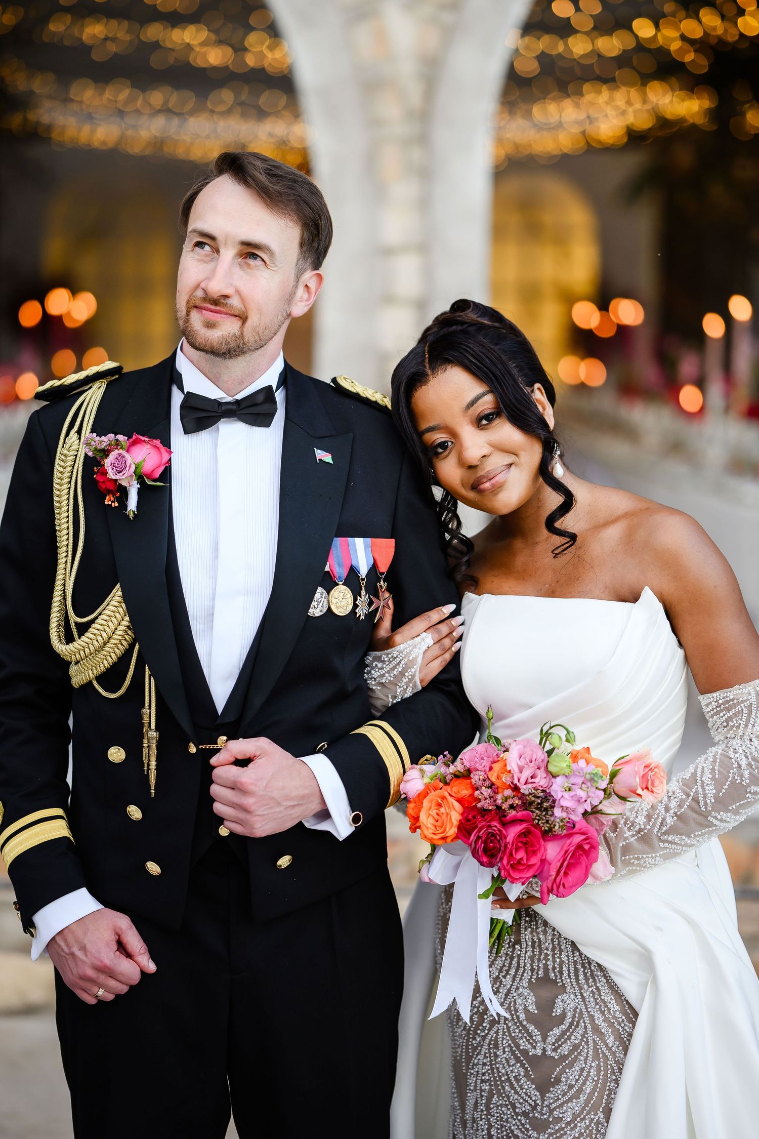 A romantic military wedding photo with a decorated officer in dress uniform and bride in white lace gown holding bright flowers.