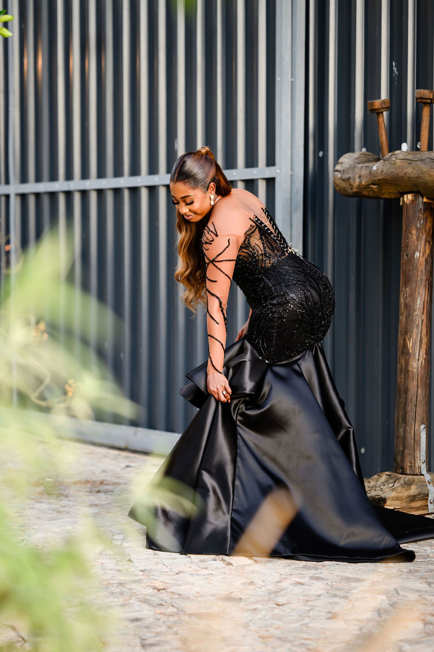 A woman in an elegant black evening gown with sheer sleeves poses gracefully against a corrugated wall background.