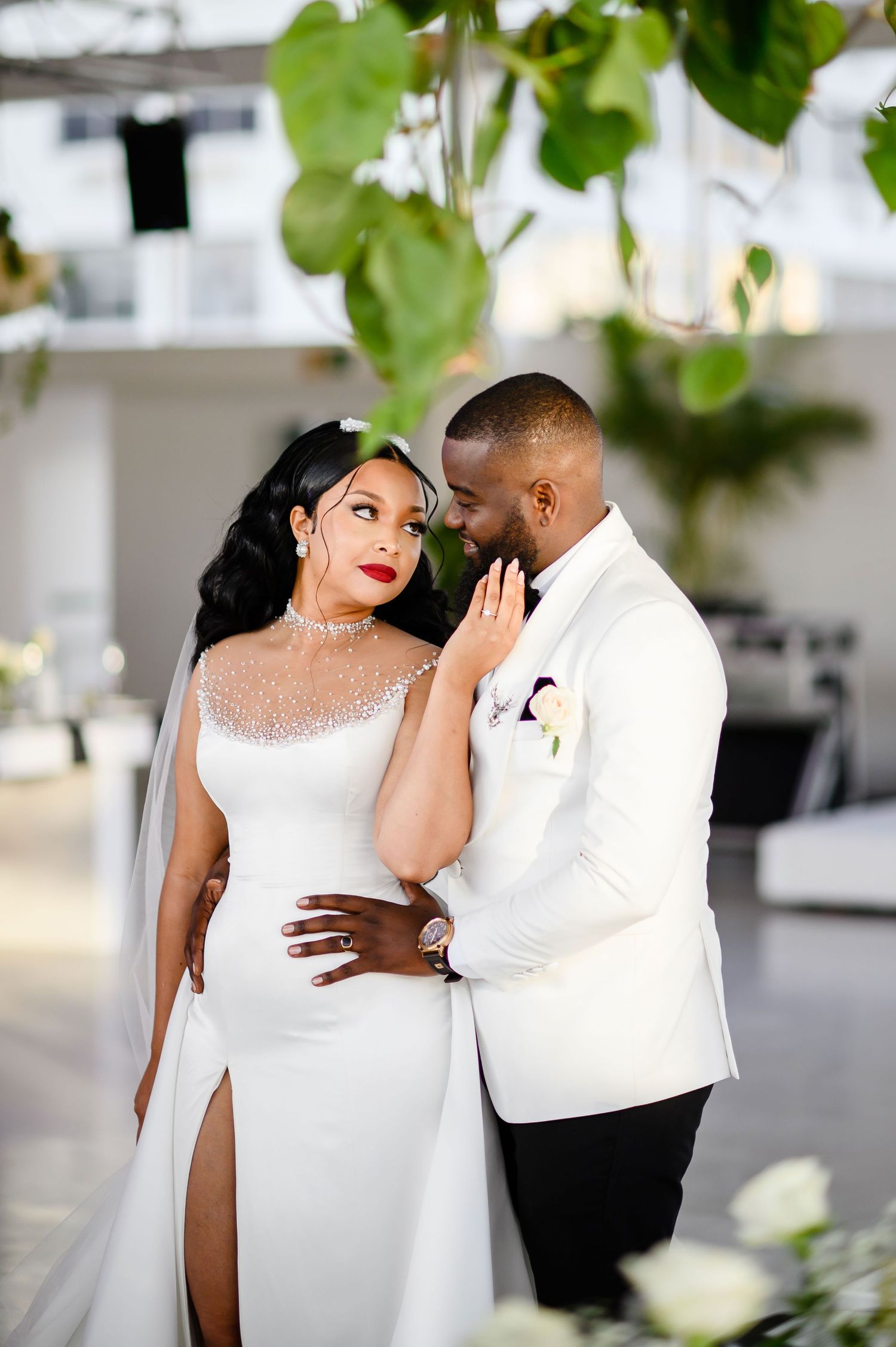 A romantic wedding photo of a couple in white attire sharing an intimate moment under green foliage.