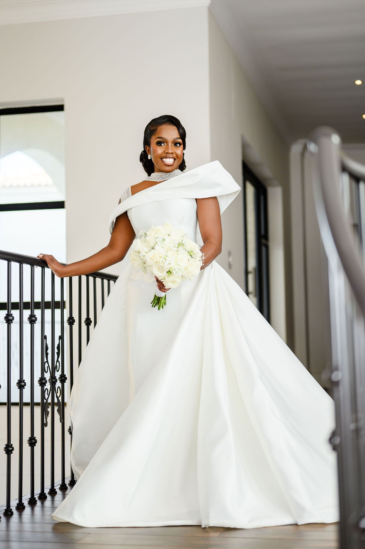Elegant off-shoulder white wedding gown with intricate bodice detailing and white floral bouquet on grand staircase.