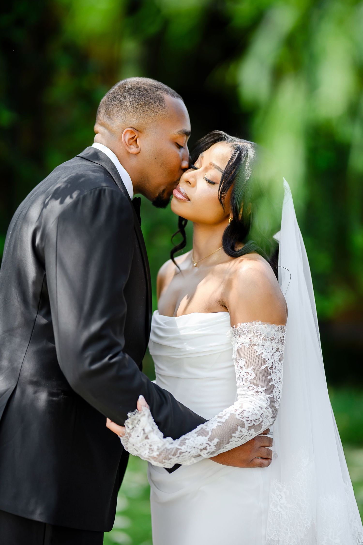 A bride in a white lace gown and groom in black suit share a romantic kiss outdoors with greenery in background.