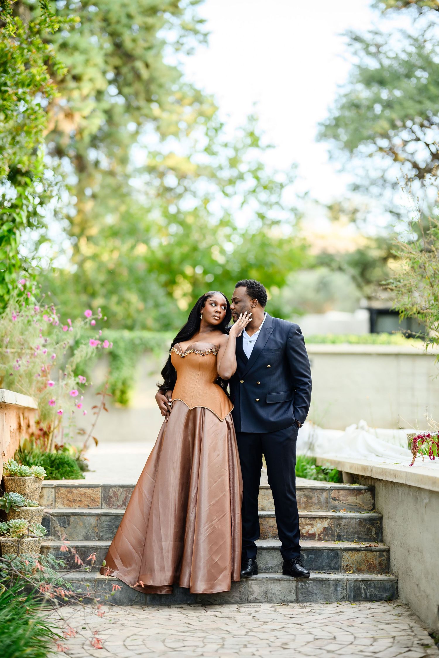 A romantic couple in formal attire poses on stone garden steps surrounded by lush greenery near a pond.