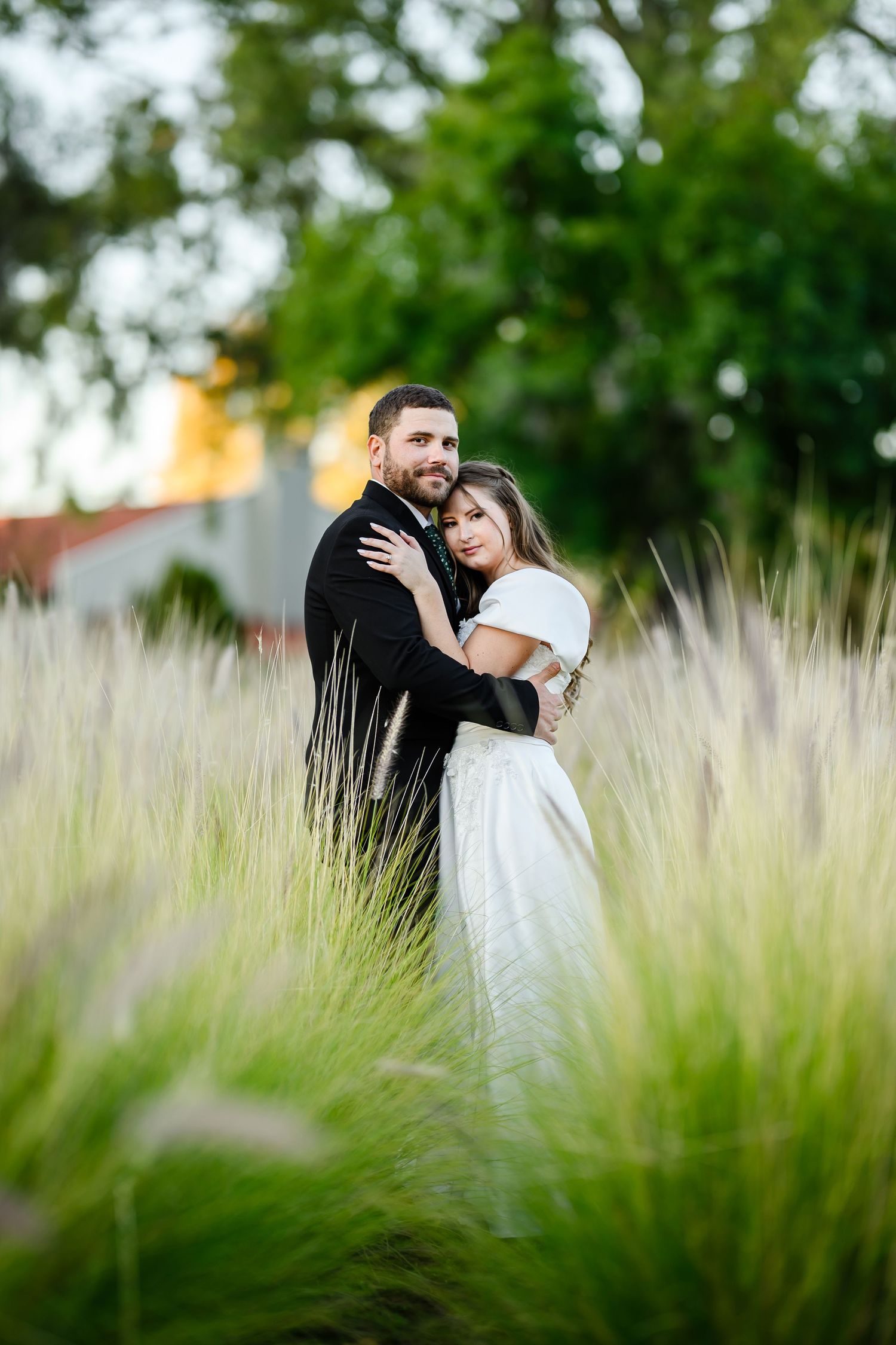 A romantic couple embraces among tall ornamental grasses in a garden setting with soft natural lighting.