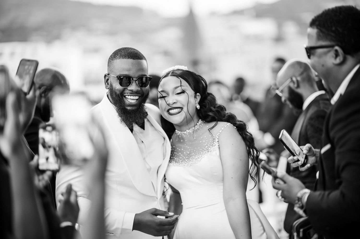 A joyful couple shares a candid moment at their outdoor wedding reception as guests surround them with phones.