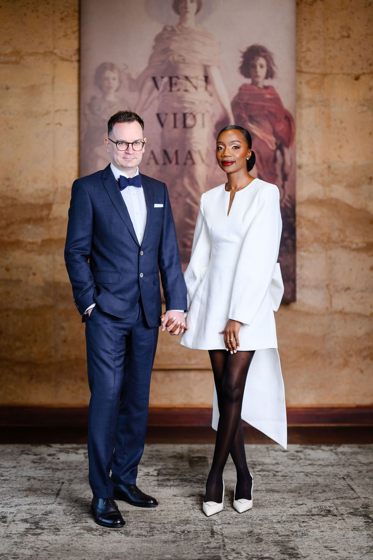 A stylish couple poses together, with one in a navy suit and bowtie and the other in an elegant white asymmetrical dress.