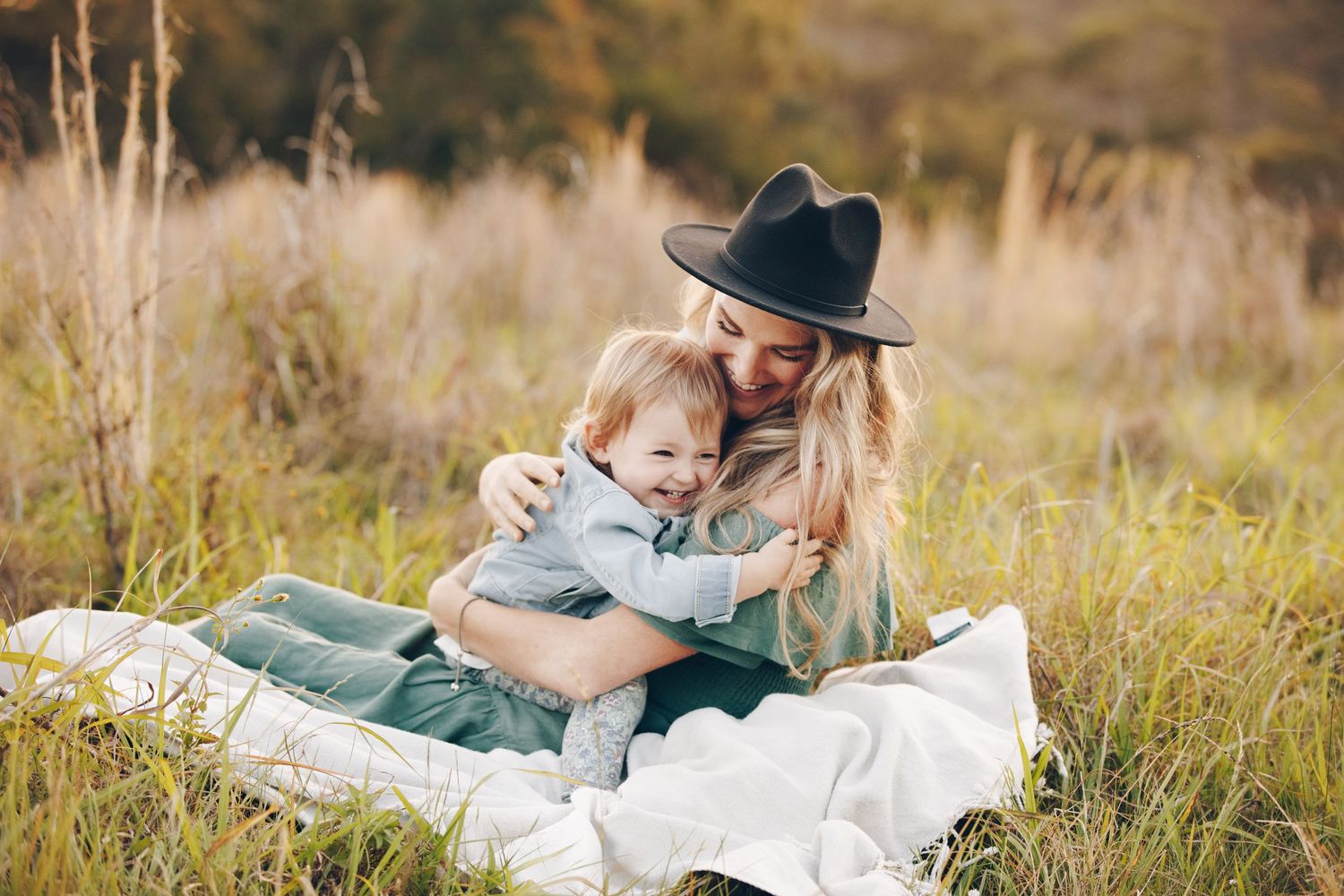 A mother in a black hat hugs her laughing toddler on a white blanket in a golden field.