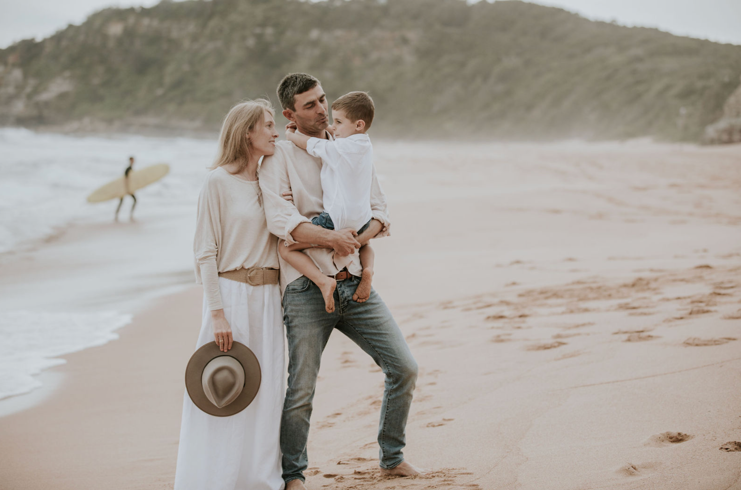 Couple holding toddler on sandy beach with surfer in background.