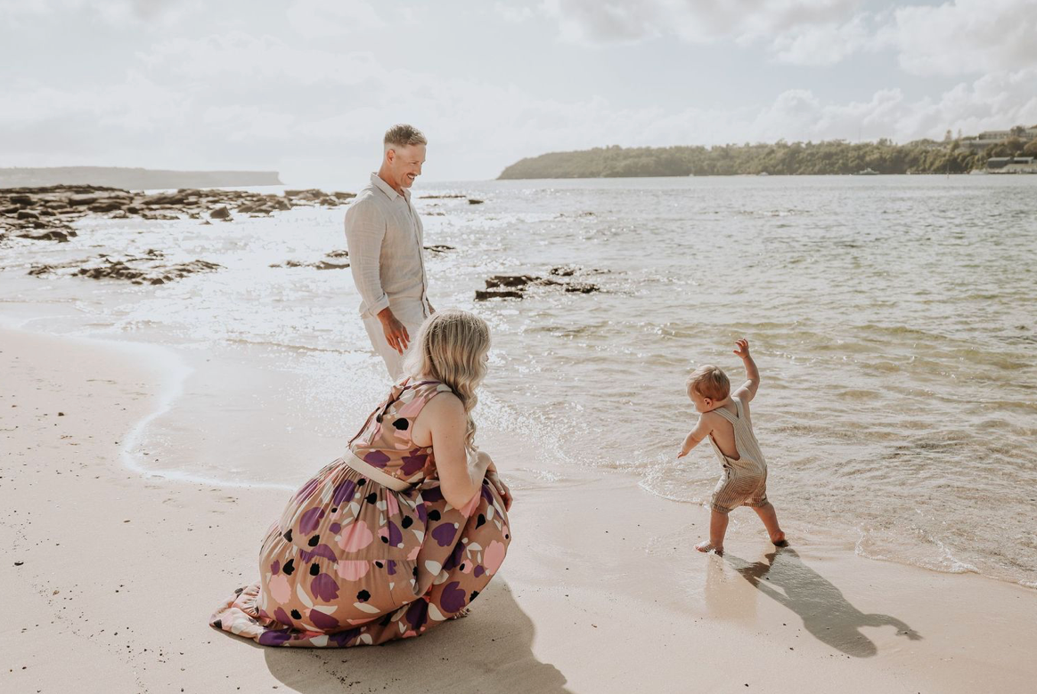 Family of three on beach shoreline, toddler splashing in waves while parents watch lovingly.