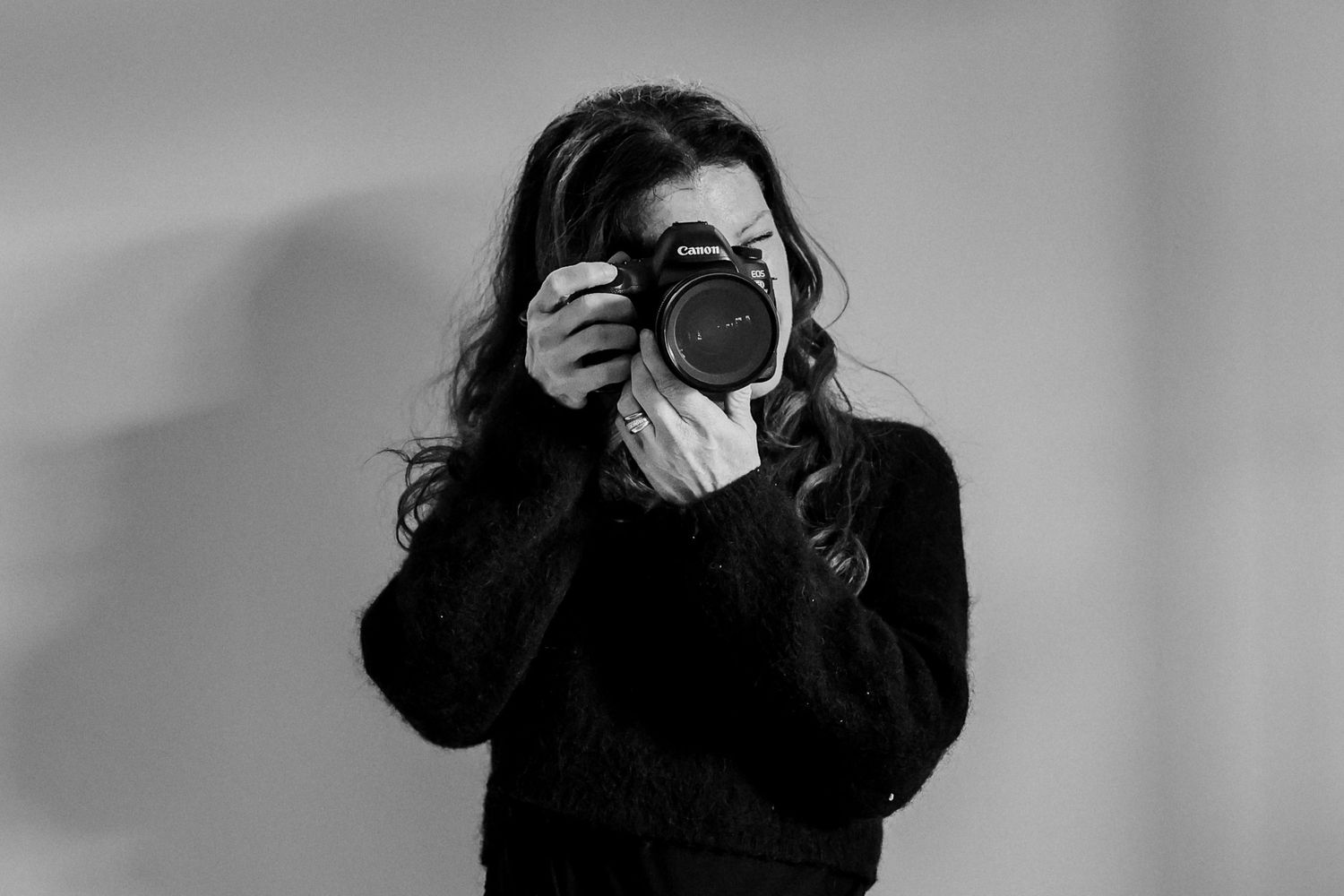 Black and white photo of a woman holding a DSLR camera up to her face, wearing a dark sweater.