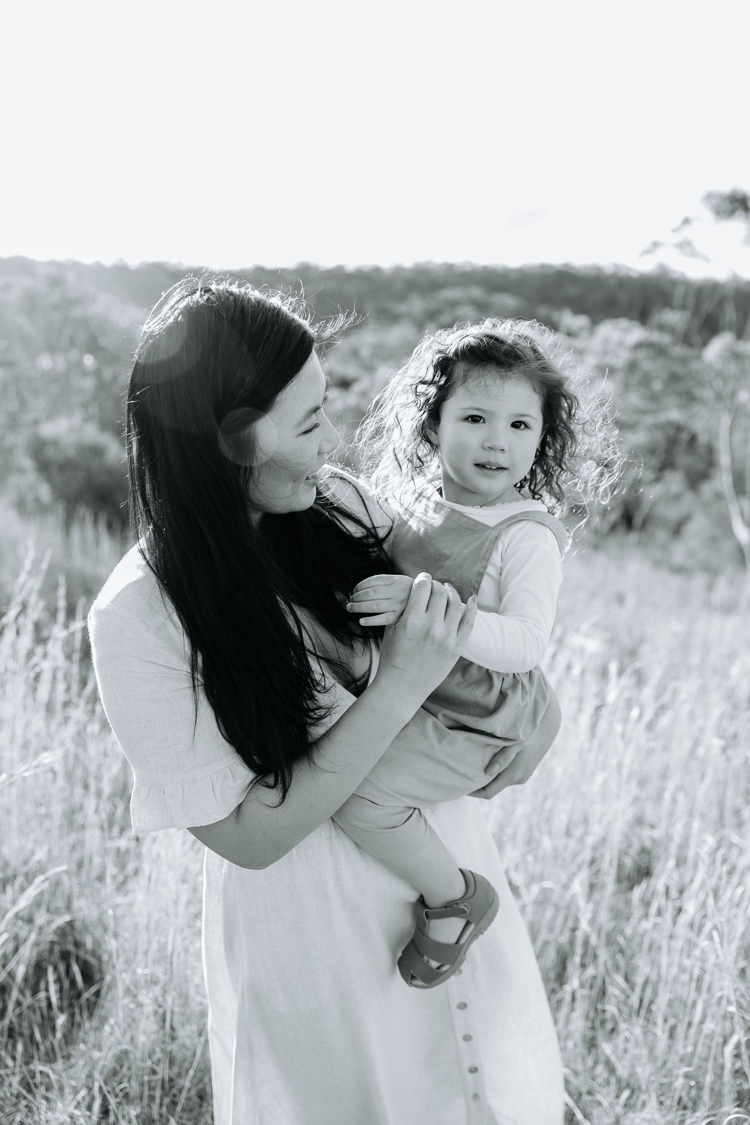 Dark-haired woman in white dress holding curly-haired toddler in field, black and white photo.