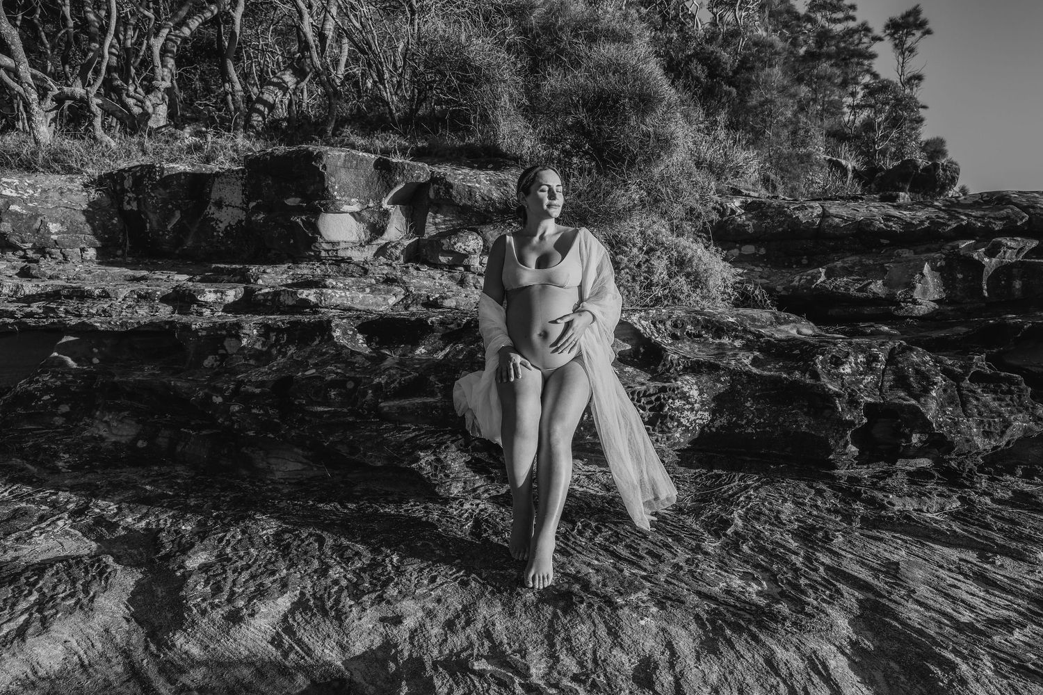 Black and white maternity photo of pregnant woman sitting on coastal rocks in sheer white robe.