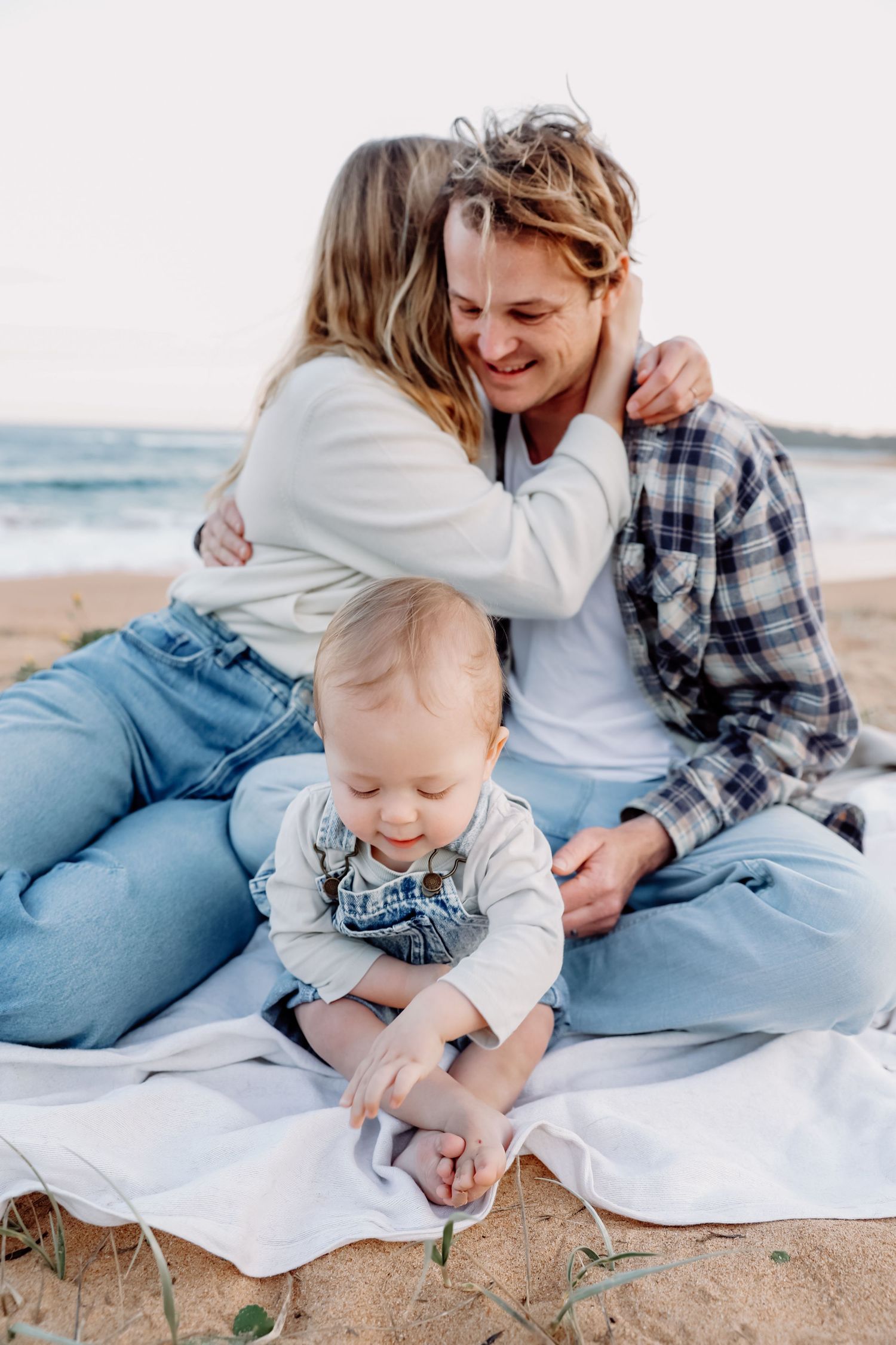 Two adults and baby sitting on blanket on beach, sharing a warm embrace by the ocean.